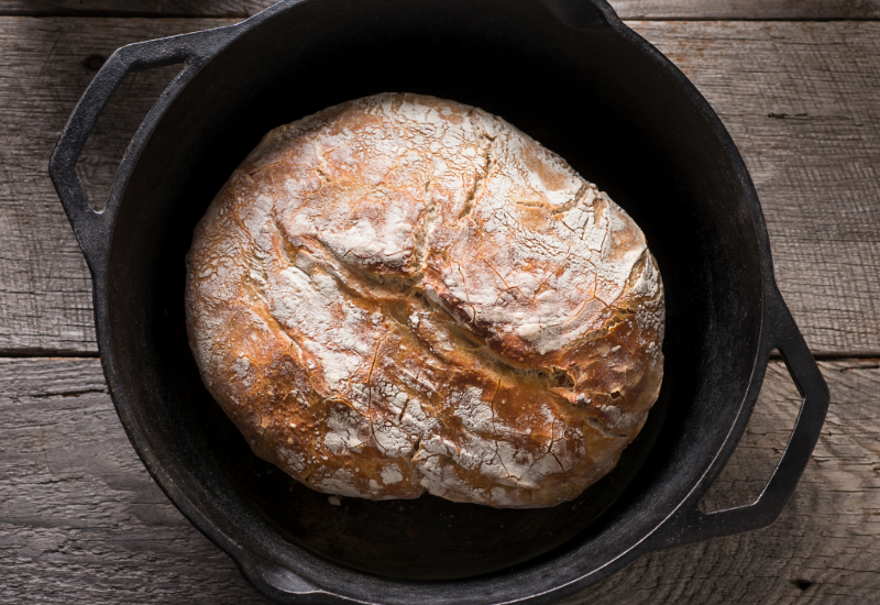 An overhead shot captures a rustic loaf of bread sitting in a seasoned cast iron pot, resting on a wooden table. The bread has a golden crust and a soft and fluffy interior, thanks to the even heat distribution of the seasoned cast iron. The pot provides a rustic and charming touch to the presentation of the bread, adding to the overall appeal of the dish. This rustic bread is perfect for any meal, whether it's served as a side, for breakfast, or as a delicious snack.