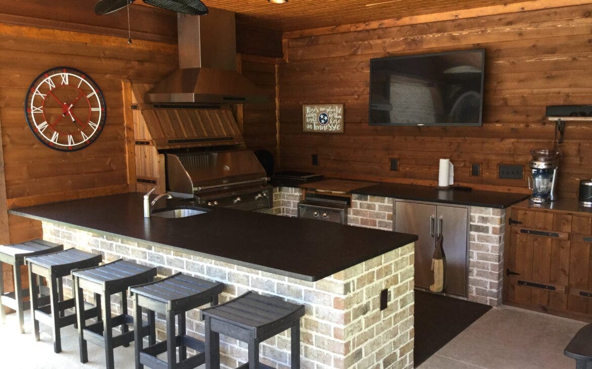 Cozy Cabin Kitchen Retreat: Proline hood keeps the air clear in this functional outdoor kitchen. Warm wood walls and brick base create a rustic charm. Ample storage and comfy seating invite conversation and lingering over meals. - Proline Range Hoods - prolinerangehoods.com 