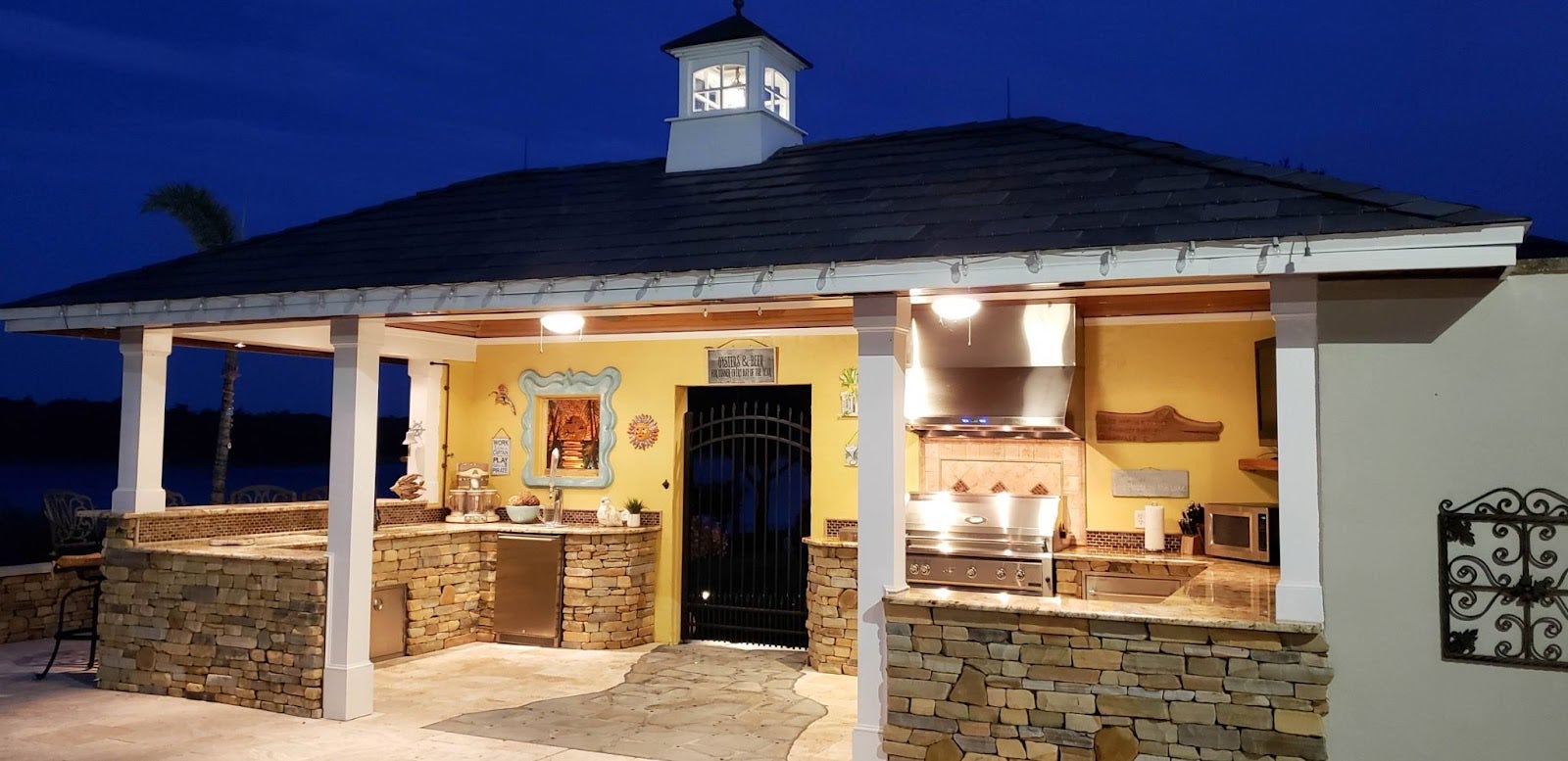 Serene Evening Kitchen: Proline hood keeps air clear for grilling under the stars in this charming outdoor kitchen. Warm lights highlight stone and yellow walls, creating a cozy space for gatherings or solo meals. - Proline Range Hoods - prolinerangehoods.com 