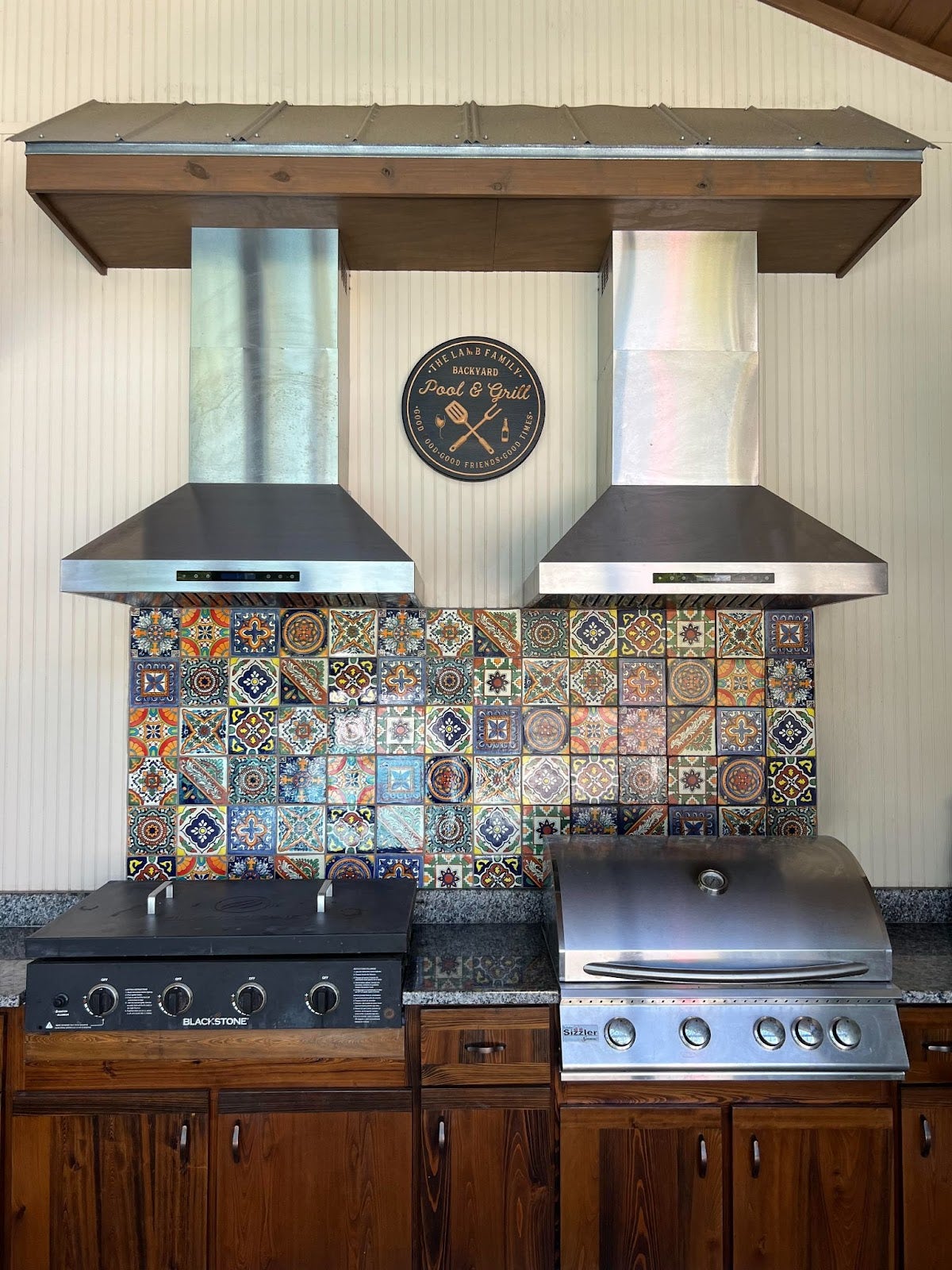Outdoor kitchen featuring a Blackstone flat-top grill and Sizzler grill with stainless steel exhaust hoods and colorful backsplash tiles - Proline Range Hoods - prolinerangehoods.com 