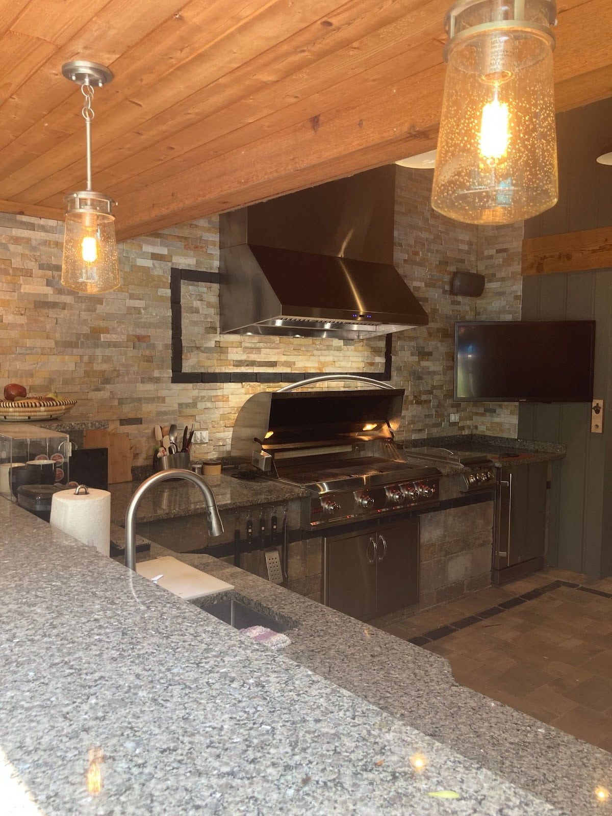 Rustic indoor kitchen with exposed wooden beams and stone wall accents, featuring a stainless steel professional grill and range hood. - Proline Range Hoods - prolinerangehoods.com 