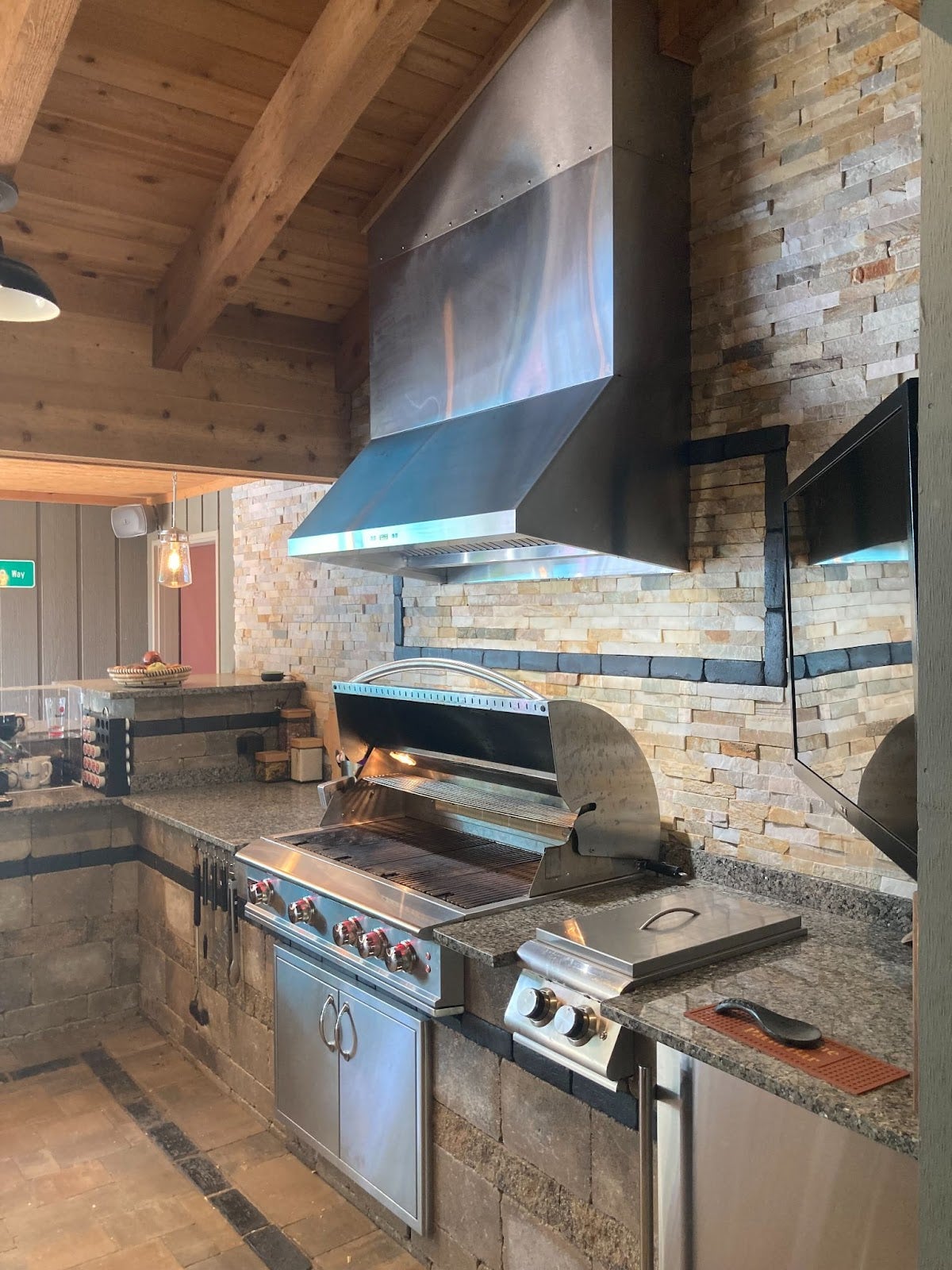 Gourmet indoor barbecue area with a high-end gas grill, prominent steel vent, granite countertops, and natural wood and stone finishes.  - Proline Range Hoods - prolinerangehoods.com 
