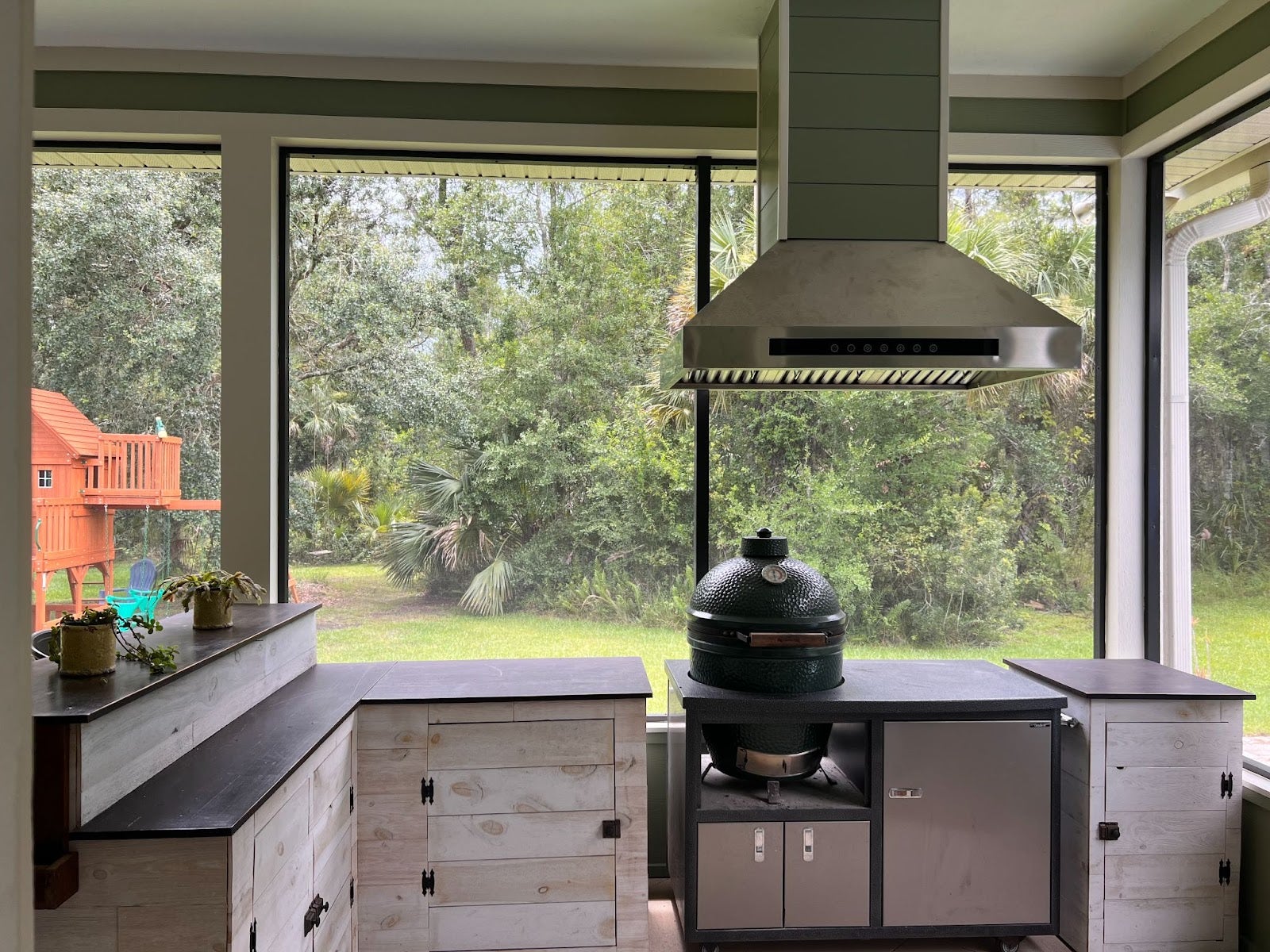 Modern outdoor grilling area featuring a green ceramic egg smoker, stainless steel vent hood, and light wood storage cabinets with a view of a backyard forest and children's play structure. - Proline Range Hoods - prolinerangehoods.com 