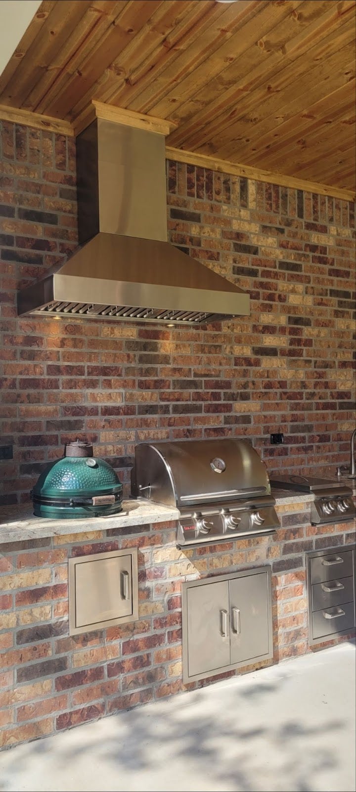 Spacious patio grilling area featuring a premium stainless steel BBQ, a Big Green Egg smoker, and a rustic brick backdrop with wooden overhead. - Proline Range Hoods - prolinerangehoods.com 