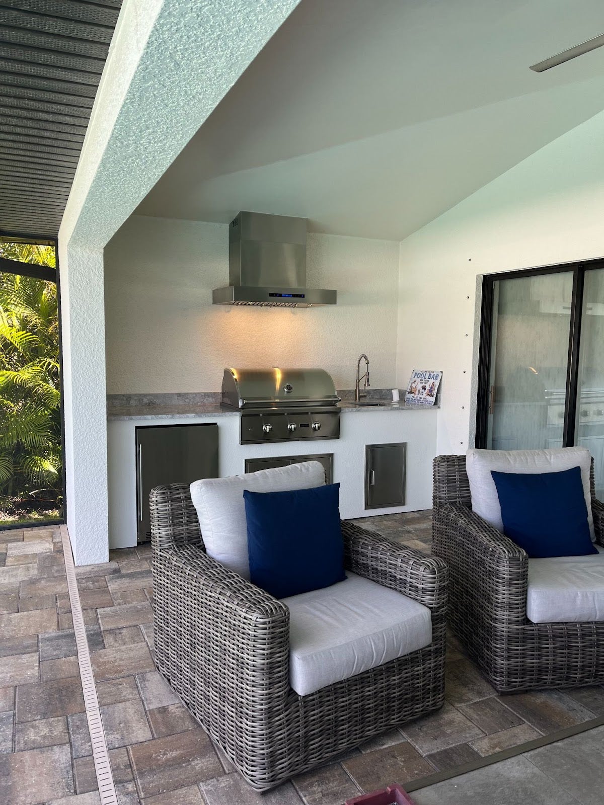 Cozy outdoor patio area with wicker armchairs and cushions, complete with a built-in barbecue  - Proline Range Hoods - prolinerangehoods.com  and stainless steel mini fridge under a modern vent hood.
