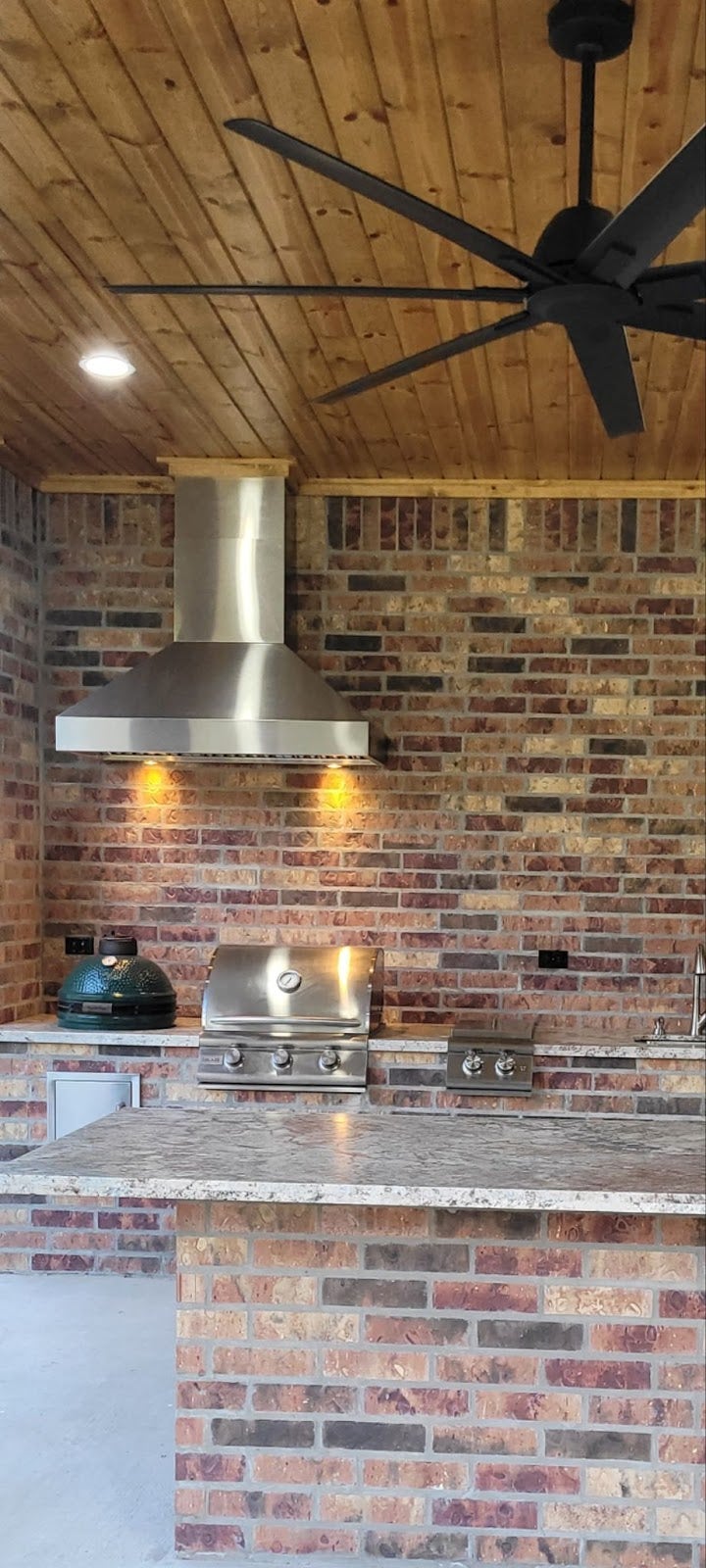 Rustic yet modern outdoor kitchen featuring a brick wall, stainless steel grill and smoker, under a wooden ceiling with a large fan. - Proline Range Hoods - prolinerangehoods.com 