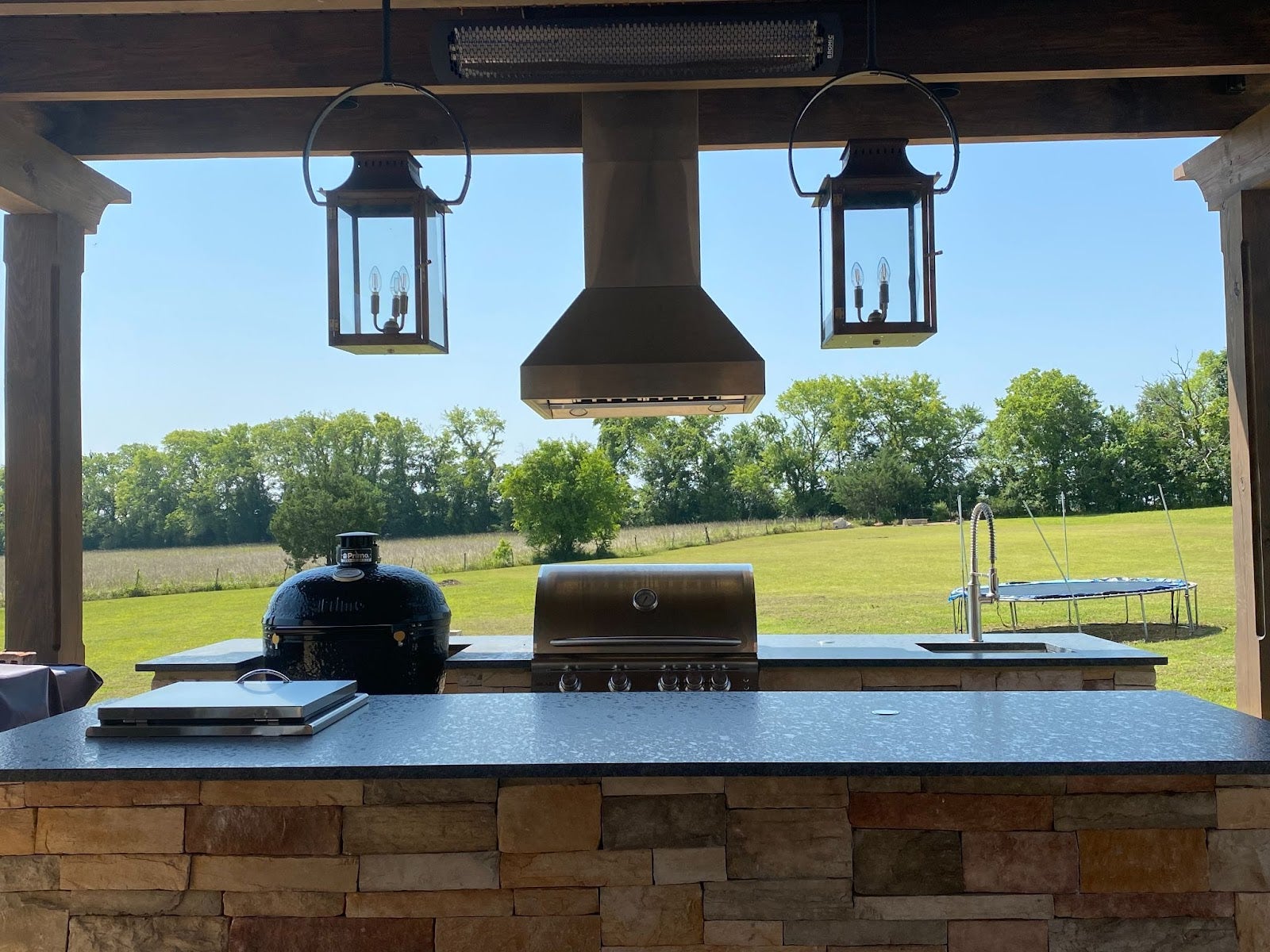 Open-air cooking area featuring a charcoal smoker, built-in gas grill, and farmhouse-style lanterns, set against the backdrop of a lush field and blue skies. - Proline Range Hoods - prolinerangehoods.com 