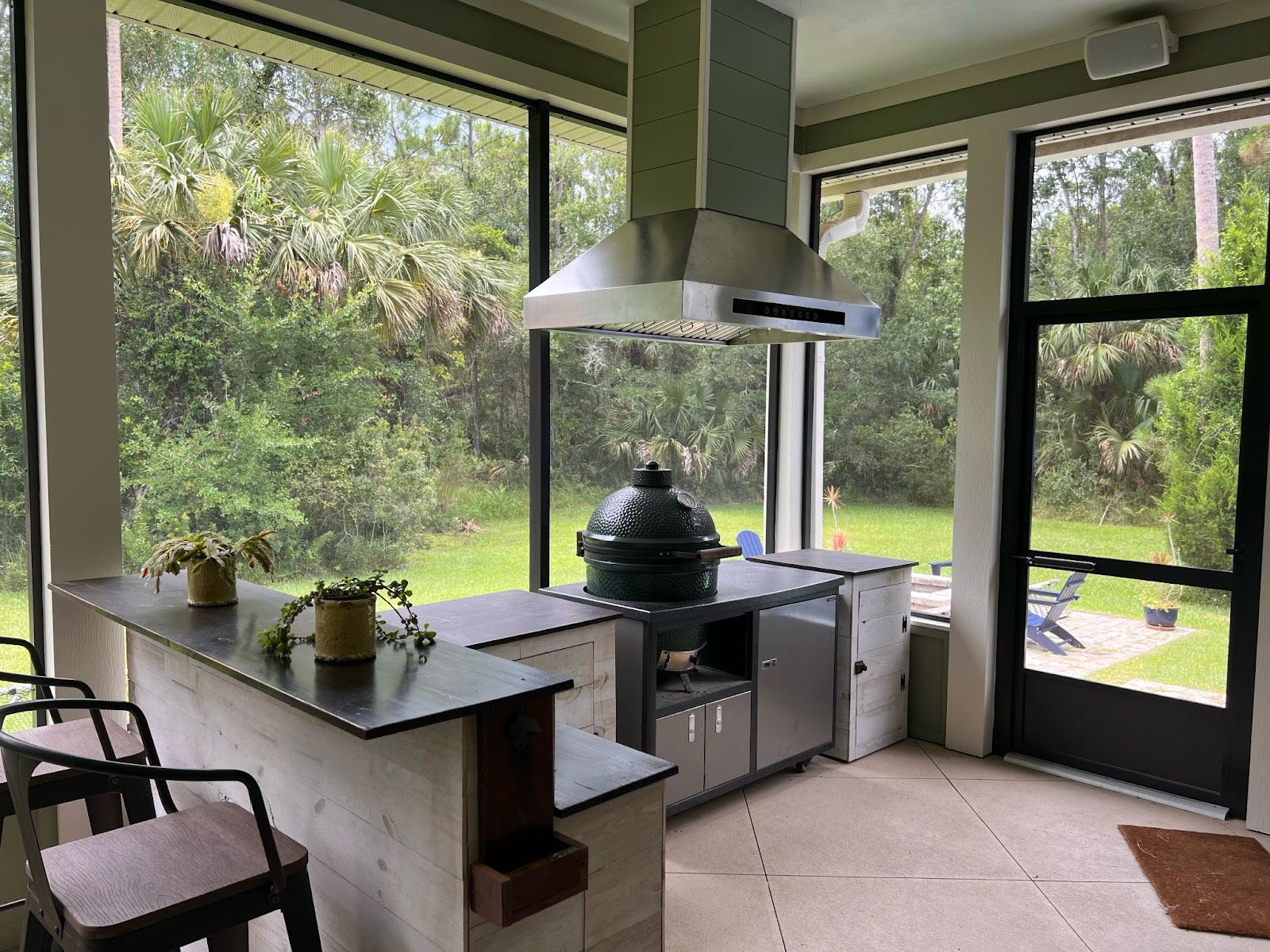 Spacious outdoor patio kitchen with a ceramic smoker grill and rustic wooden cabinetry set against a backdrop of lush greenery and a wooden playset. proline range hoo in an outdoor setting