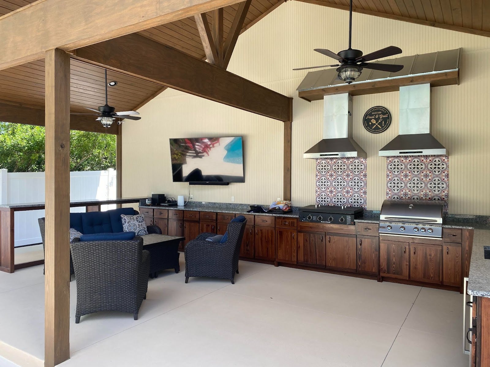 Expansive covered outdoor kitchen with dark wicker furniture, wood cabinetry, and decorative tile backsplash under a vaulted ceiling with fans two proline outdoor vent hoods over a grill and a blackstone - Proline Range Hoods - prolinerangehoods.com 