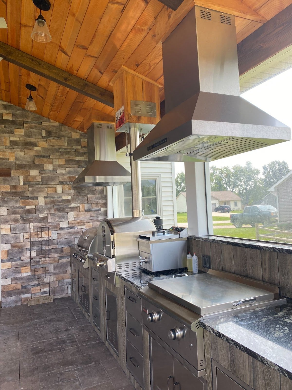 Sophisticated backyard kitchen setup featuring a built-in barbecue, smoker, and storage cabinets with a view of neighborhood homes and clear skies. - Proline Range Hoods - prolinerangehoods.com 