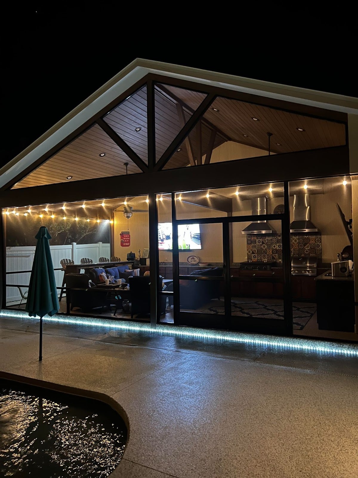 Evening atmosphere in a sheltered outdoor entertainment area with LED lighting, a poolside view, and an open-concept kitchen visible in the background. - Proline Range Hoods - prolinerangehoods.com 