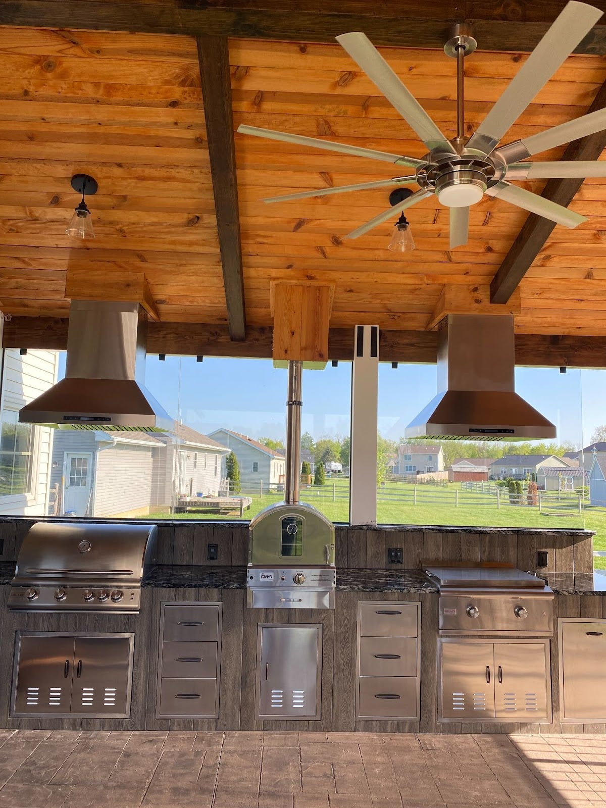 Spacious outdoor kitchen with stainless steel appliances, Proline range hoods, wooden cabinets, and a large ceiling fan, set against a suburban landscape. - Proline Range Hoods - prolinerangehoods.com 