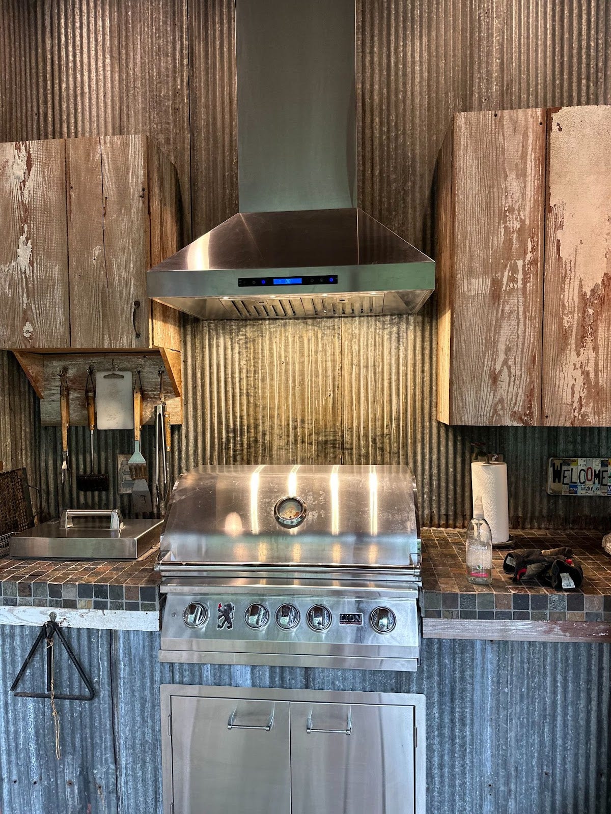 Industrial-style outdoor kitchen with stainless steel grill and range hood, against a distressed wooden and corrugated metal backdrop. - Proline Range Hoods - prolinerangehoods.com 