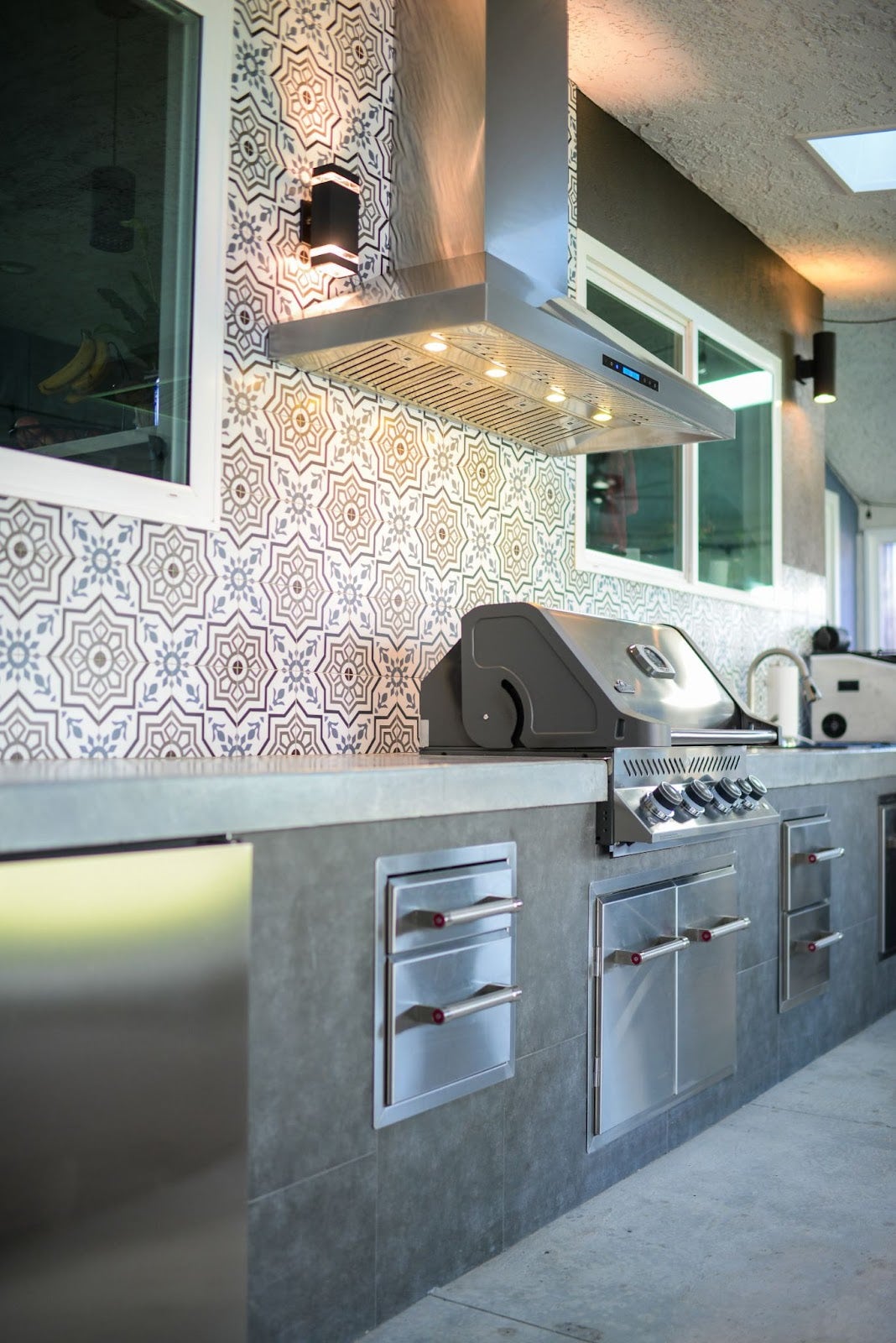 Chic Outdoor Kitchen: Bold patterned tiles and a Proline hood create a modern vibe in this outdoor kitchen. Stainless steel hood offers ventilation for smoke-free grilling. Perfect for entertaining with modern appliances and ambient lighting.  - Proline Range Hoods - prolinerangehoods.com  