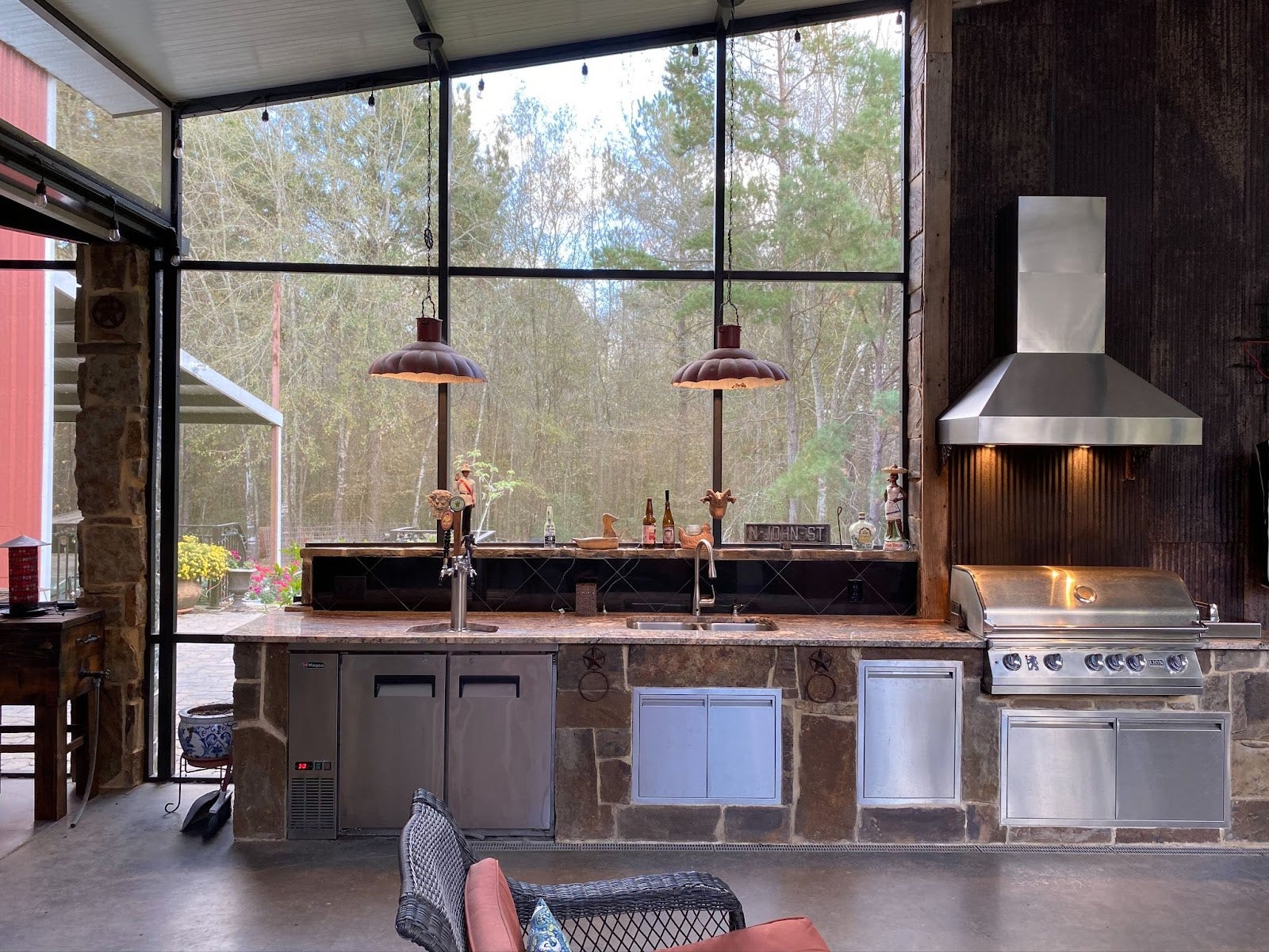 A semi-outdoor kitchen combining industrial style with rustic elements. Stainless steel range hood contrasts with wood and stone. Large screened windows offer a view of the outdoors. Spacious layout with ambient lighting is ideal for gatherings. - Proline Range Hoods - prolinerangehoods.com 