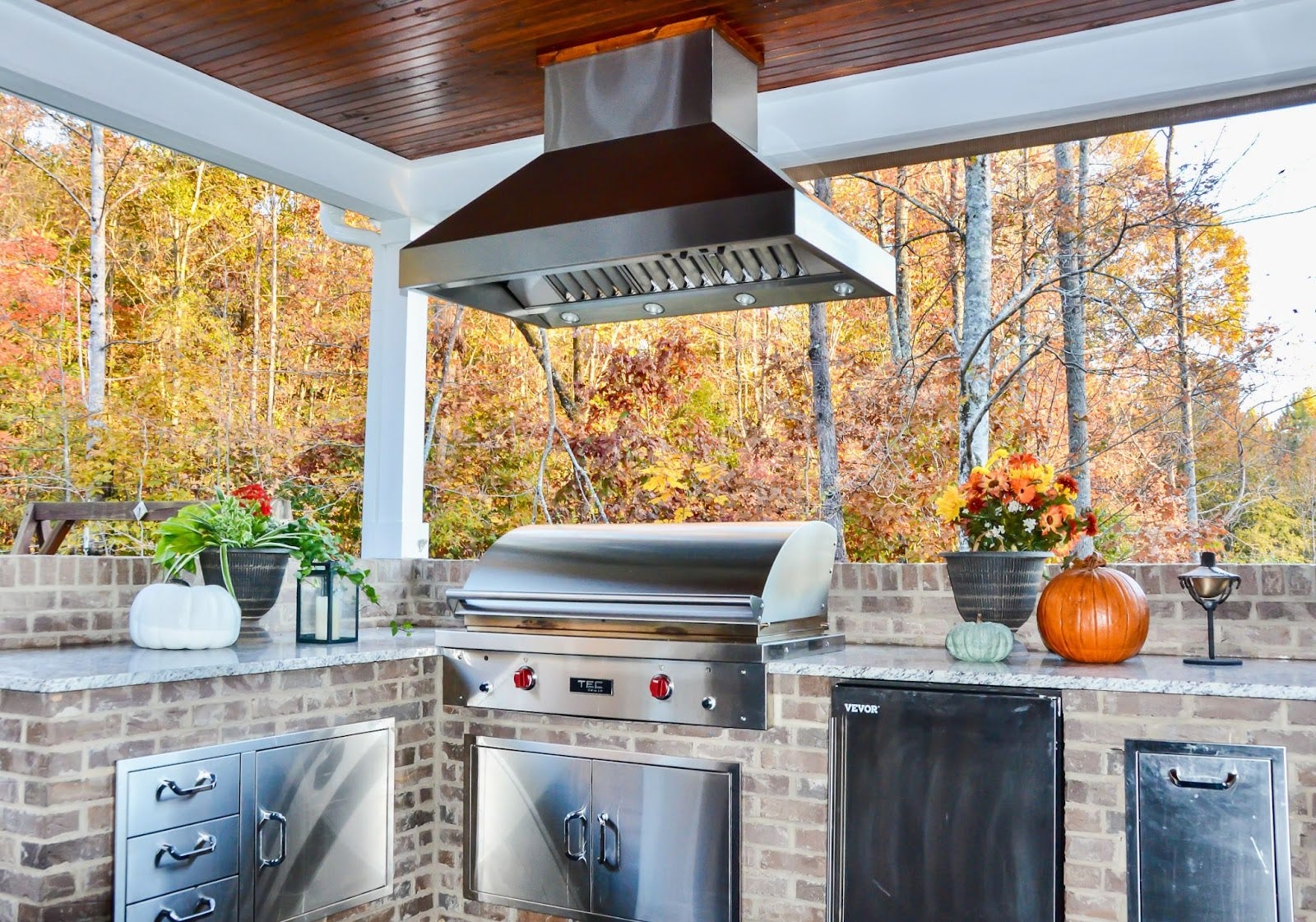 Autumn Kitchen Oasis: Proline hood adds a modern touch to this rustic outdoor kitchen with brickwork. Fall foliage creates a colorful backdrop. Seasonal decor complements the natural beauty. Perfect for grilling and enjoying the harvest season. - Proline Range Hoods - prolinerangehoods.com 