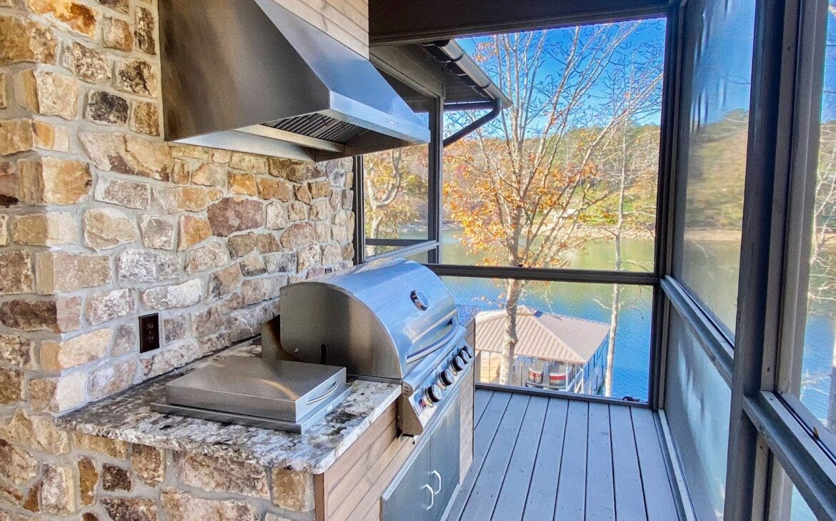 Modern open-air kitchen featuring a professional grill, range hood, and stone countertops set against a stone wall backdrop on a lake. - Proline Range Hoods - prolinerangehoods.com 