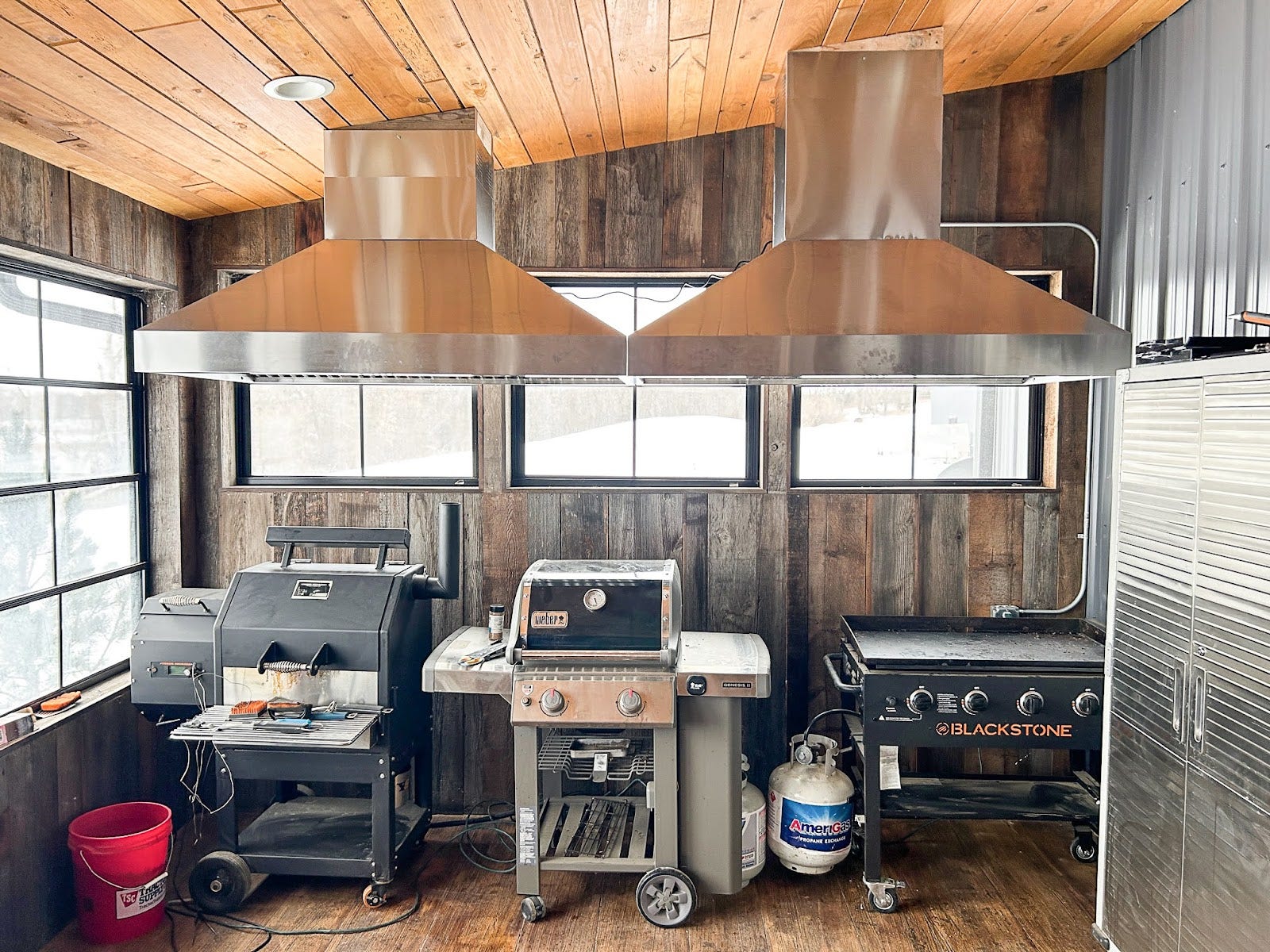 Industrial Rustic Kitchen: Raw wood walls and dual PLJI 103 range hoods create a bold, industrial-rustic atmosphere. The wide stainless steel hoods handle serious cooking with ease, venting smoke from grills under the large windows filled with natural light. Embrace the joy of cooking in this unique outdoor space! - Proline Range Hoods - prolinerangehoods.com 