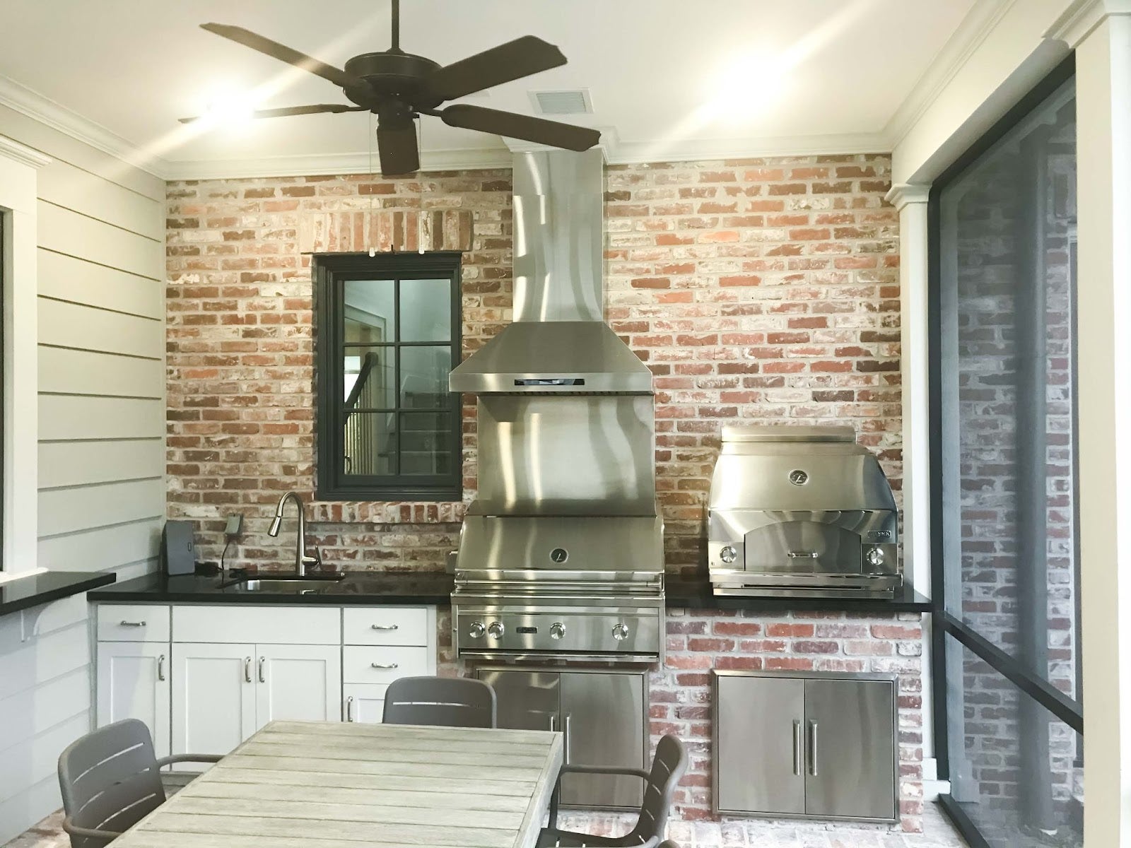 Rustic Modern Grilling Station: Exposed brick walls meet modern stainless steel range hood in this functional outdoor kitchen. The spacious grill and ample storage make it perfect for family BBQs. A ceiling fan keeps things cool, while the window offers a view of the outdoors. Perfect for creating lasting memories! - Proline Range Hoods - prolinerangehoods.com 