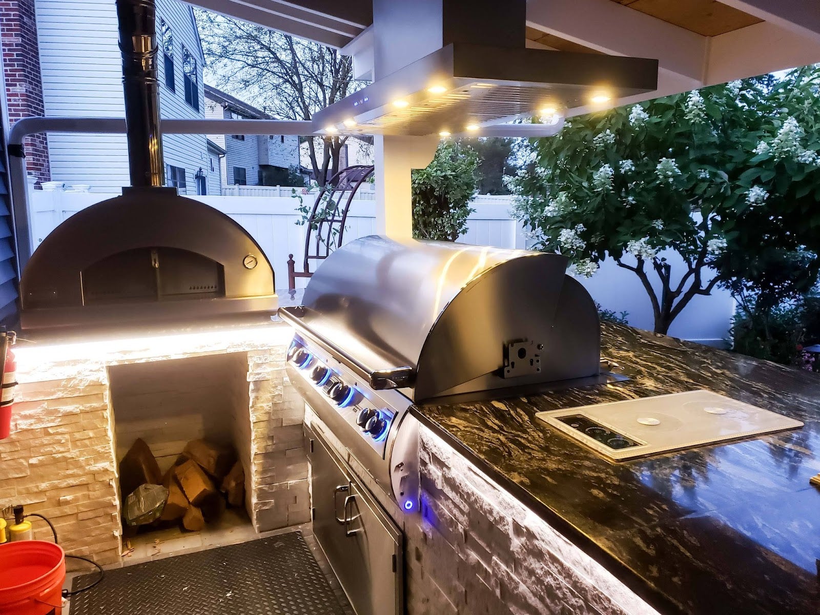 Alfresco Kitchen with Pizza Oven: This inviting outdoor kitchen features a matte black pizza oven, stainless steel grill, and Proline vent hood. Under-cabinet lighting creates a warm ambiance. Stone veneer and wood accents blend with the greenery for a sophisticated, earthy feel. - Proline Range Hoods - prolinerangehoods.com 
