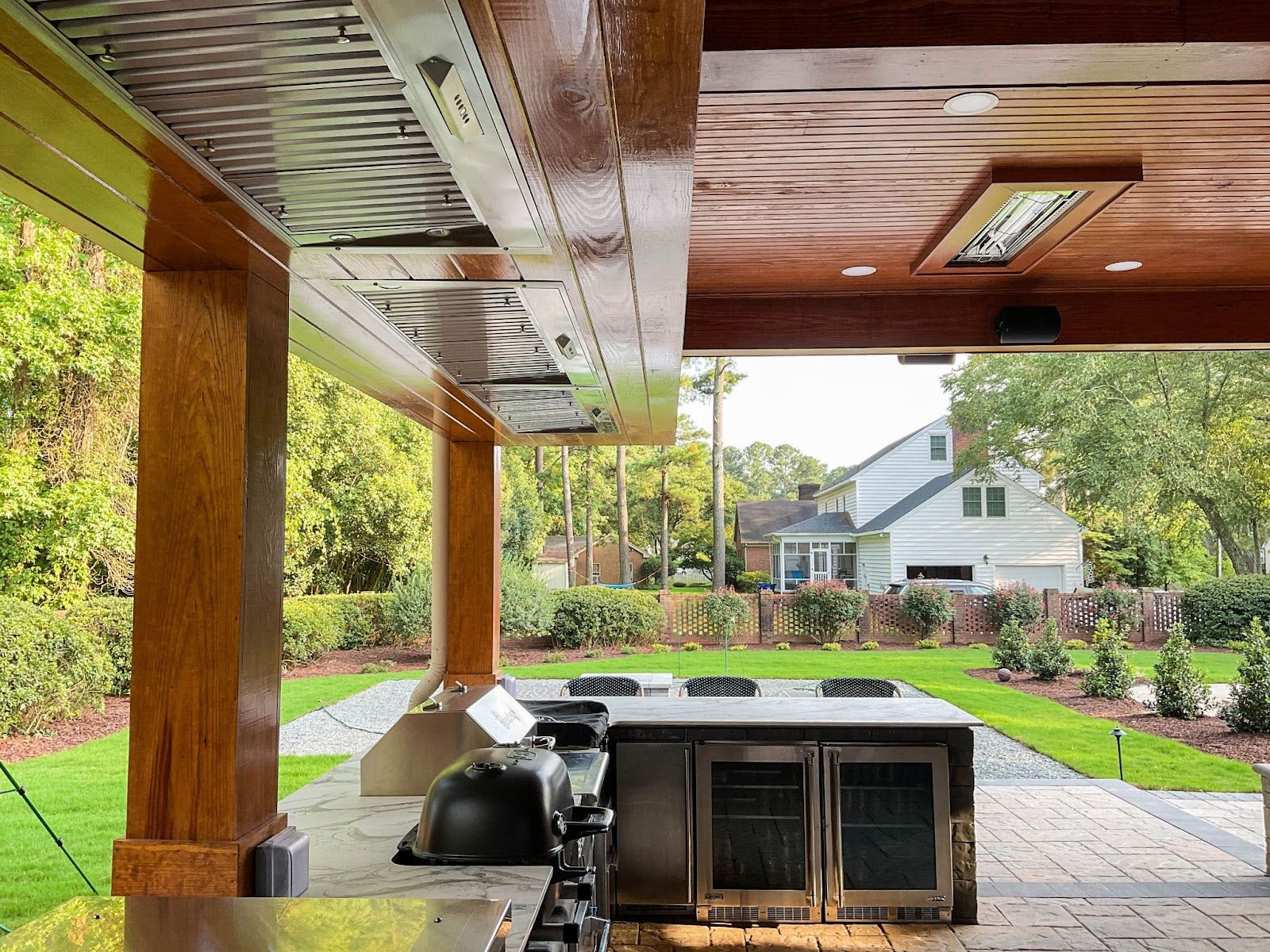 Chef's Outdoor Kitchen: Rustic meets modern! Sheltered patio with wood beams & stainless steel appliances. Multiple range hoods keep the air clear. Skylight bathes the space in light. Landscaped garden completes the view. - Proline Range Hoods - prolinerangehoods.com 