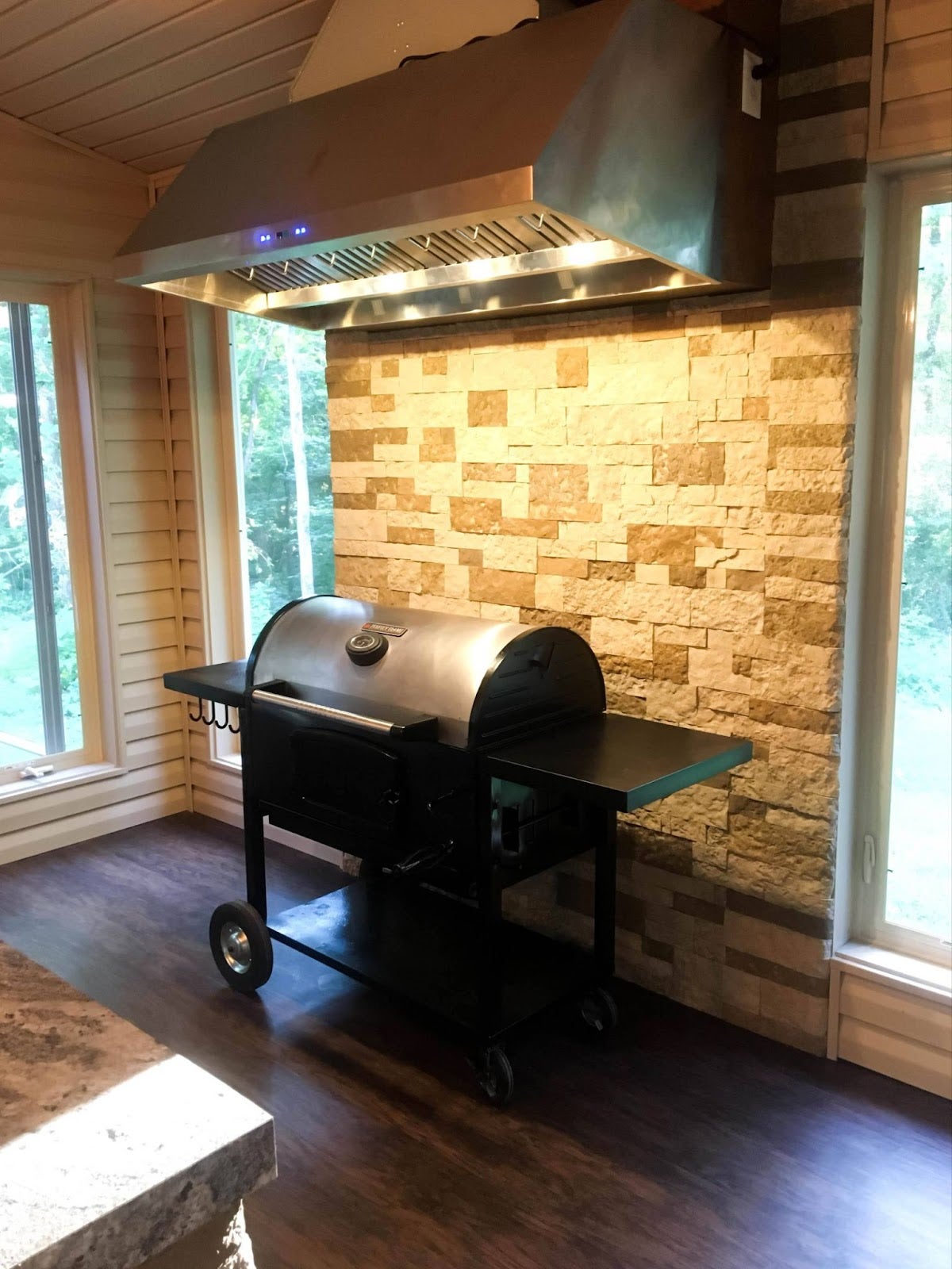 Functional Indoor Grilling Station: Black pellet grill with Proline PLFW 832 range hood in a versatile indoor/outdoor space. Brushed stainless steel hood complements the wood and stone, with task lighting and large windows for a bright ambiance. - Proline Range Hoods - prolinerangehoods.com 