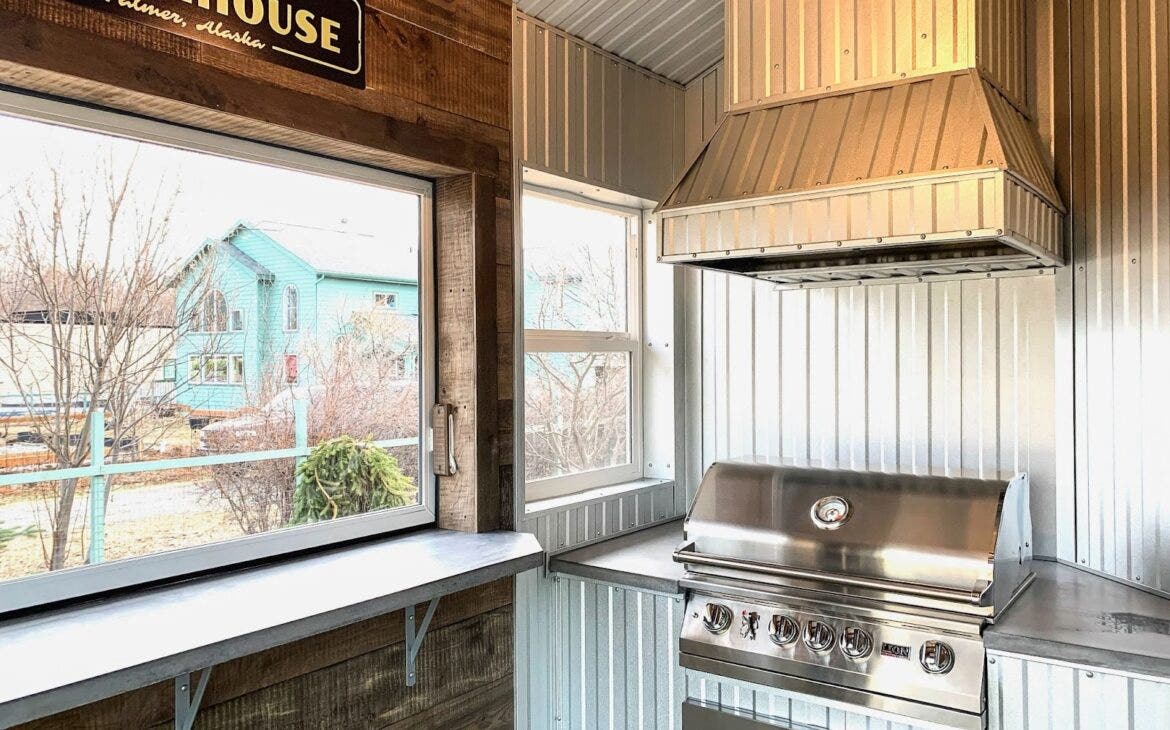 Industrial Chic Alaskan Kitchen: Stainless steel Proline hood and corrugated metal walls create an industrial vibe in this outdoor kitchen. Warm wood accents and soft lighting add Alaskan charm. Perfect for smoky barbecues and relaxed gatherings. - Proline Range Hoods - prolinerangehoods.com 