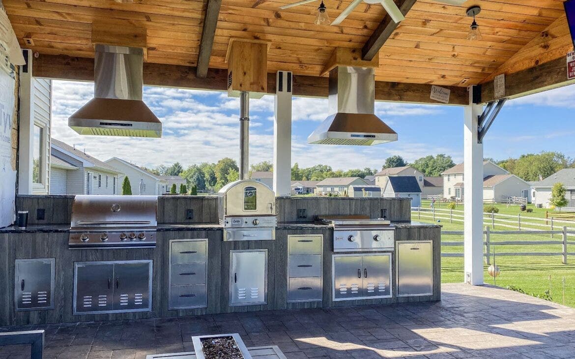 Heartland Grill Symphony: Three Proline hoods ensure smokeless grilling in this rustic outdoor kitchen. Wooden beams and a gentle breeze create a relaxing atmosphere. Perfect for celebrating the outdoors and enjoying gourmet meals. - Proline Range Hoods - prolinerangehoods.com 