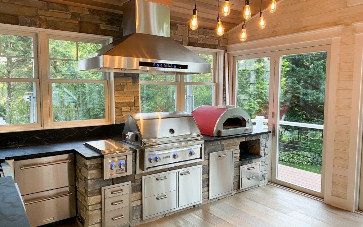 Rustic-Luxe Kitchen with Pizza Oven: Proline PLJW 102 hood complements stainless steel appliances in this kitchen. Stone counters, warm wood tones, and a pizza oven create a welcoming ambiance. Edison bulbs and expansive windows add charm and natural light. - Proline Range Hoods - prolinerangehoods.com 