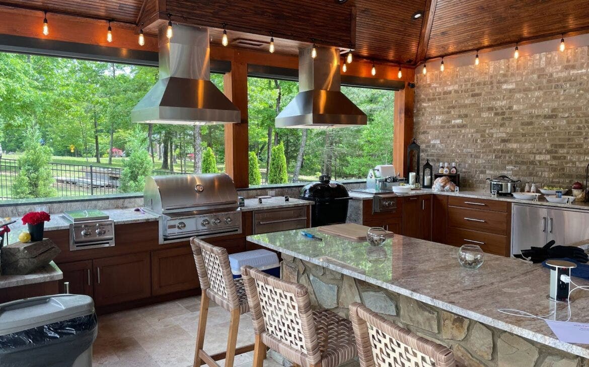 Charismatic Outdoor Kitchen: Twin Proline PLJI 103 hoods vent smoke for the twin grills in this mountain lodge-inspired kitchen. Wood ceiling, stone accents, and patterned stools create a playful ambiance. Lush greenery adds a natural touch. - Proline Range Hoods - prolinerangehoods.com 