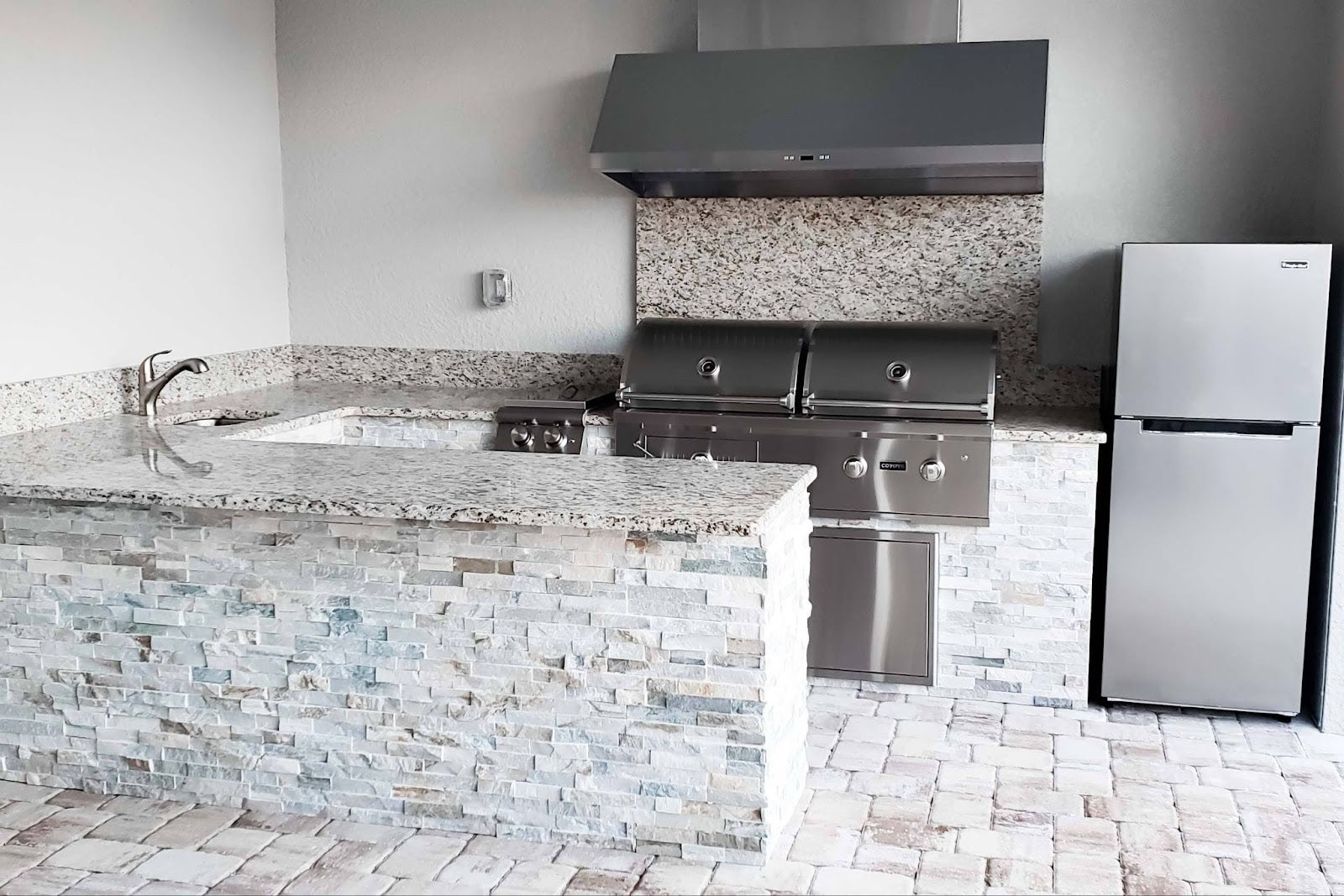 Relaxed Outdoor Kitchen: Stainless steel grill on a stone island with granite countertops. Spacious layout for easy grilling and entertaining. Smoke-clearing range hood and nearby fridge keep things convenient. Perfect for creating memories! - Proline Range Hoods - prolinerangehoods.com 
