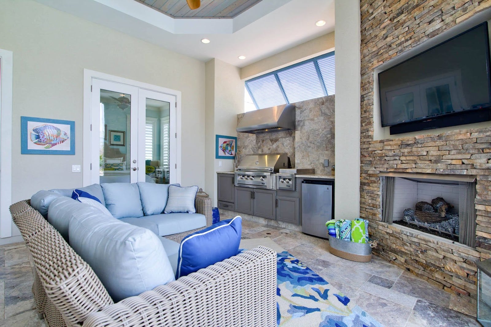 Open living room with plush blue sectional sofa, stone fireplace, gourmet kitchen featuring stainless steel appliances and bar seating, sliding glass doors to outdoor space, and seaside decor accents. - prolinerangehoods.com