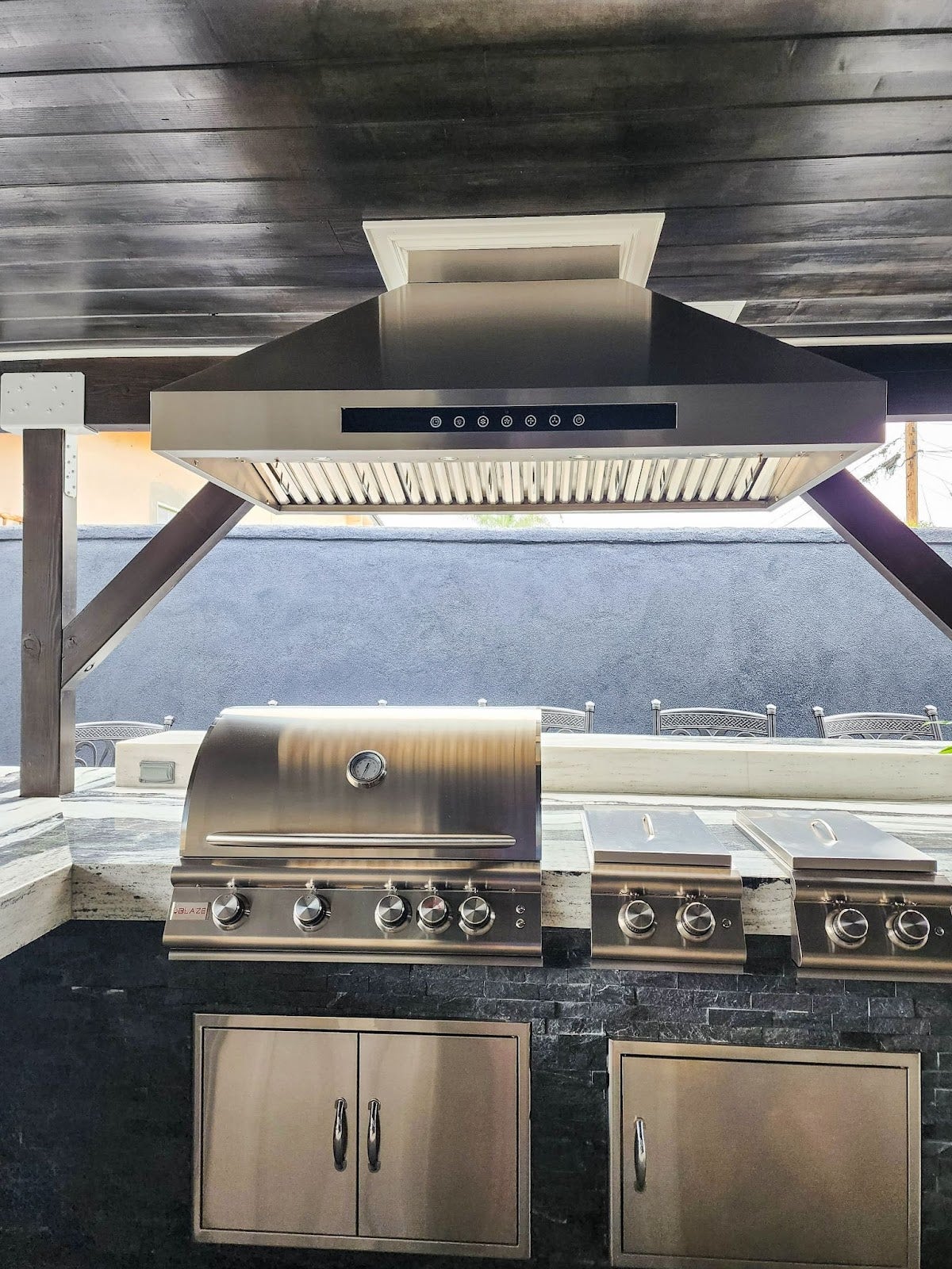 Contemporary outdoor kitchen with a stainless steel BBQ grill and extractor hood, set against a dark stone wall under a stained wooden ceiling. - Proline Range Hoods - prolinerangehoods.com 