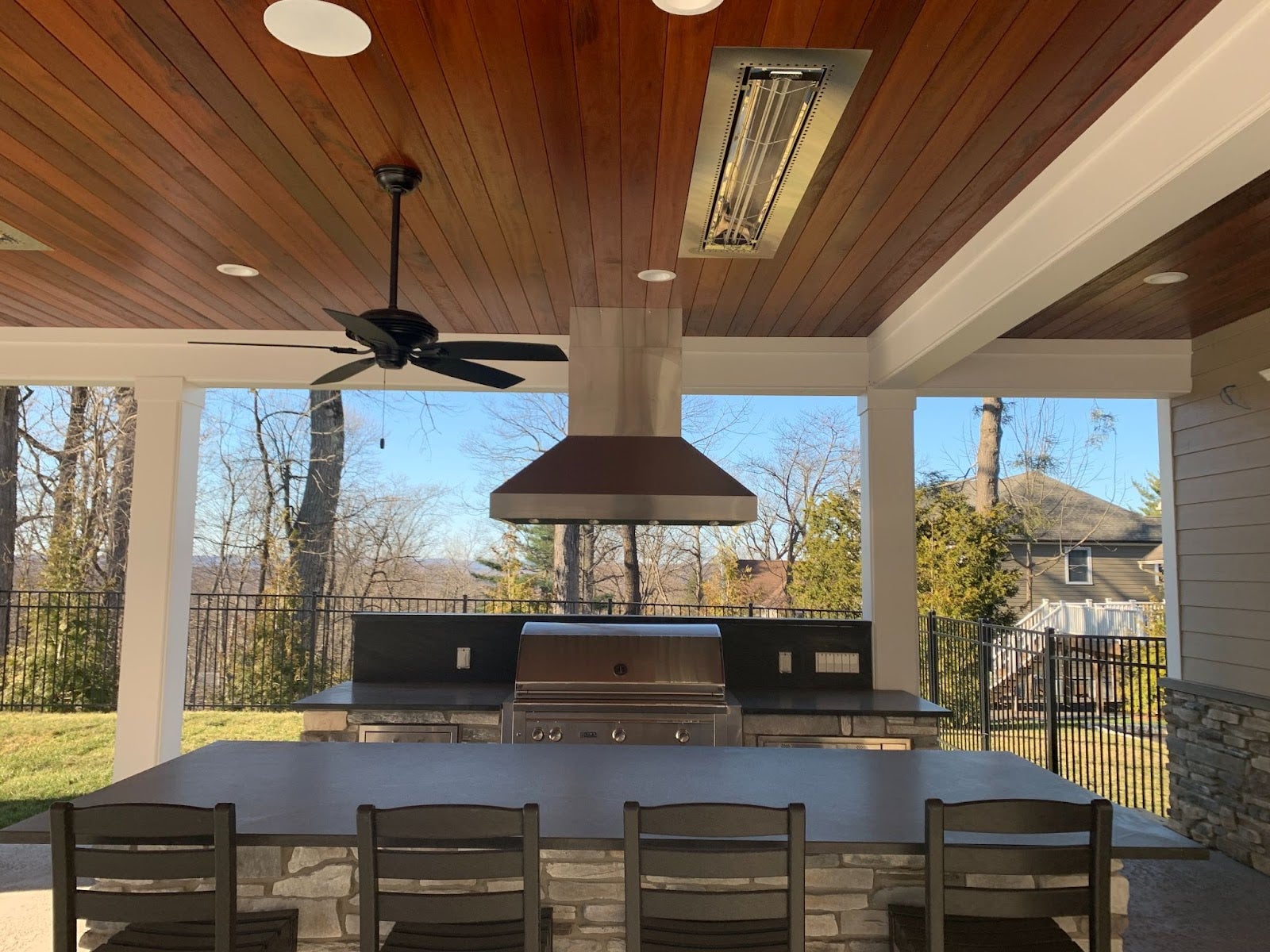 Contemporary outdoor covered patio with a wooden ceiling, featuring a large dining table and a built-in barbecue grill. - Proline Range Hoods - prolinerangehoods.com 