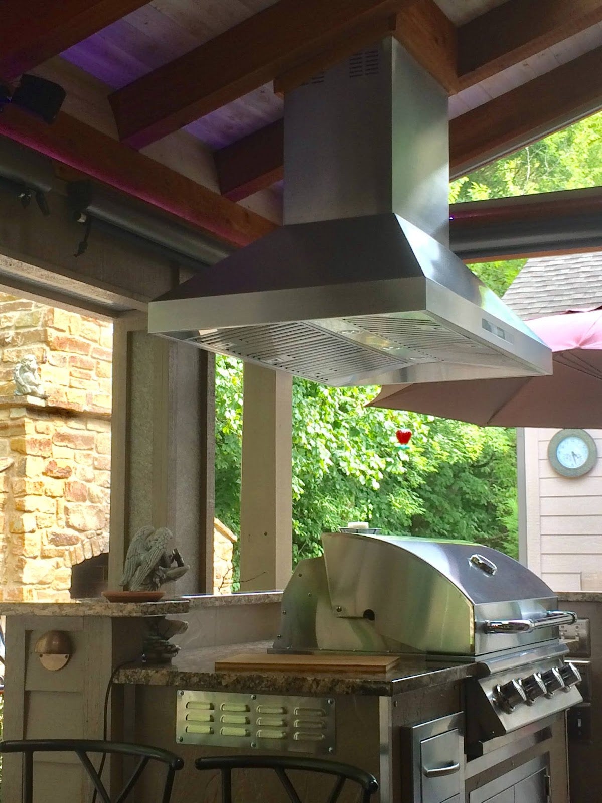 Outdoor kitchen setup with stainless steel grill under a large vent hood on a patio with exposed wooden beams. - Proline Range Hoods - prolinerangehoods.com 