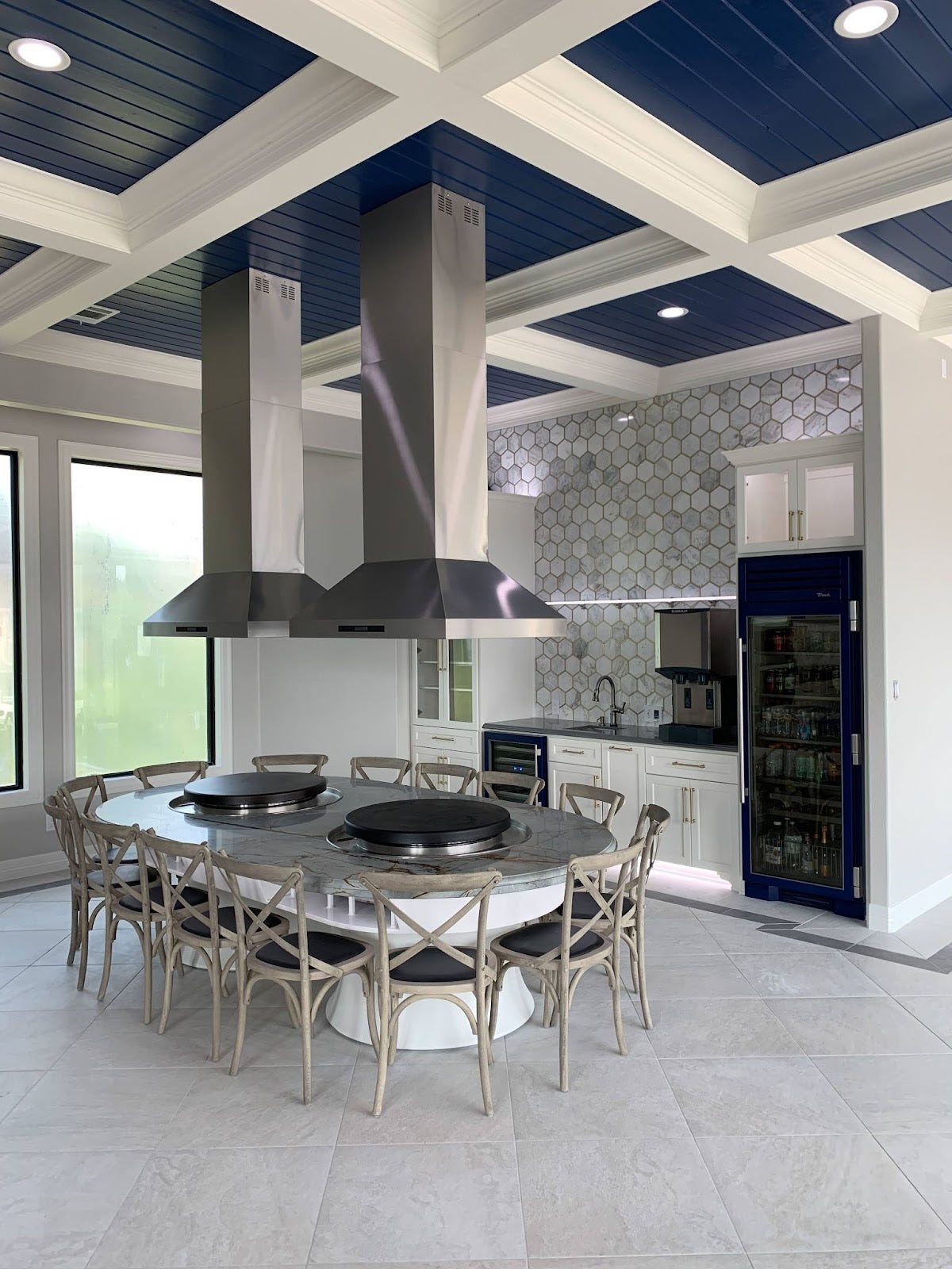 Spacious modern kitchen featuring two large extractor hoods, a central round dining table with a cooktop, and contrasting navy and white decor. - Proline Range Hoods - prolinerangehoods.com 