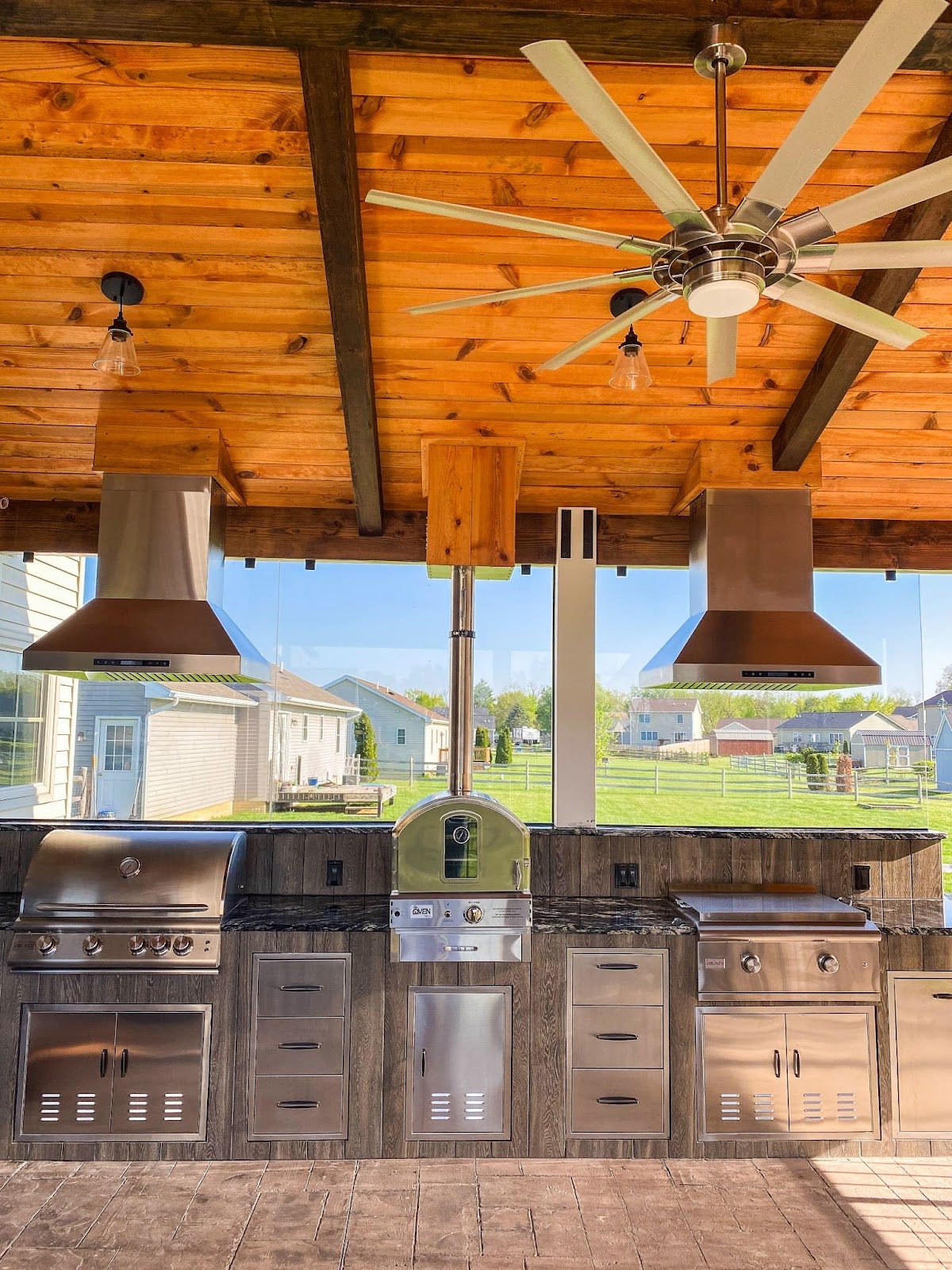 Stylish outdoor culinary setup with wood-paneled ceiling, featuring premium grilling equipment, ventilation hoods, and a view of a residential neighborhood. - Proline Range Hoods - prolinerangehoods.com 