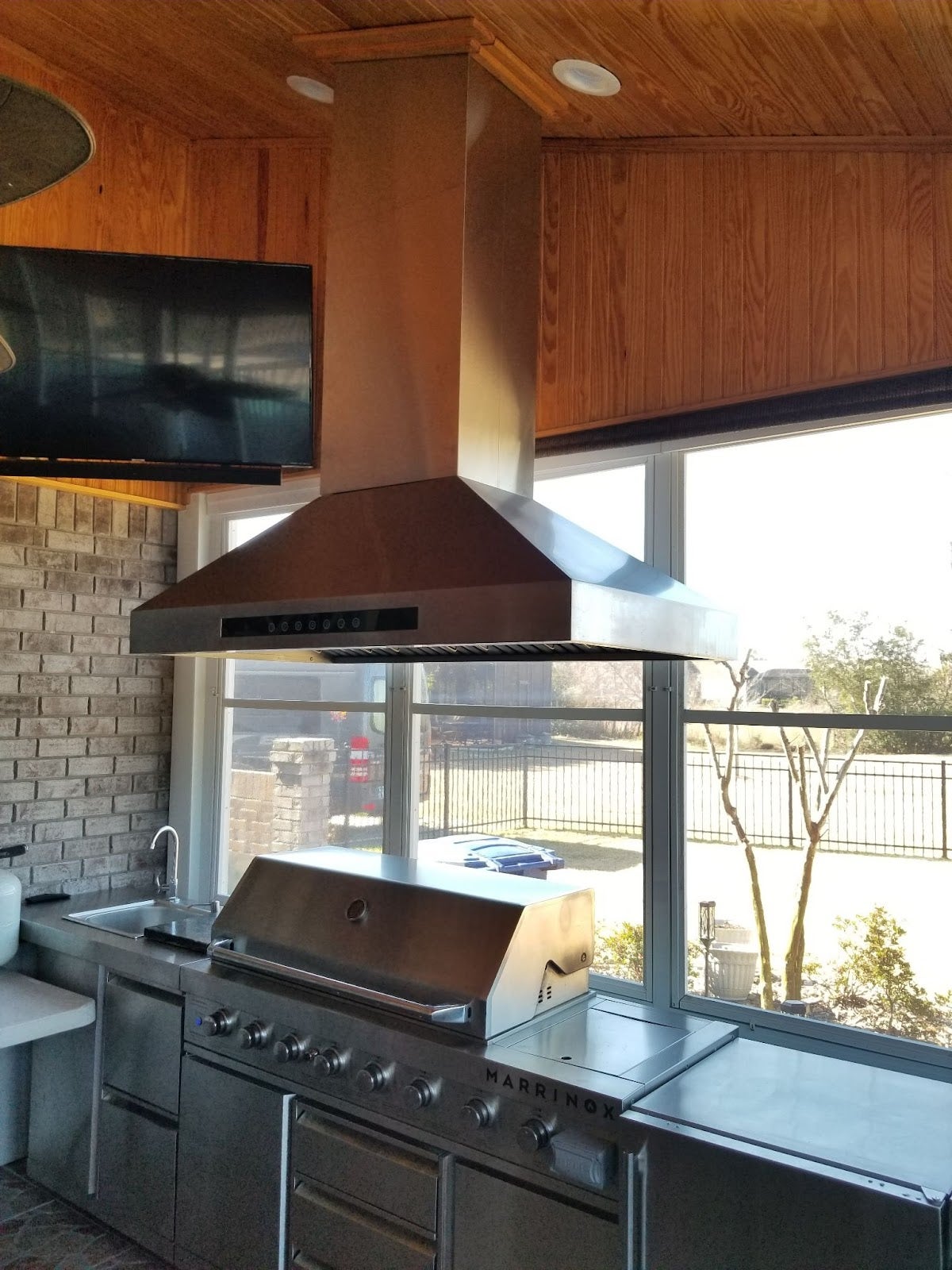 Inviting indoor patio grill area featuring a large stainless steel barbecue, extractor fan, and wooden interior details, with a view of the outdoors. - Proline Range Hoods - prolinerangehoods.com 