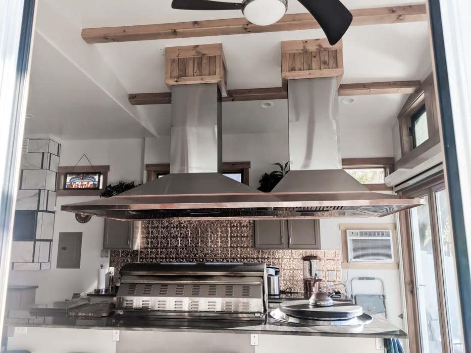 Large commercial-style kitchen featuring dual ventilation hoods over a professional cooktop, set in a room with wooden ceiling accents and contemporary design. - Proline Range Hoods - prolinerangehoods.com 