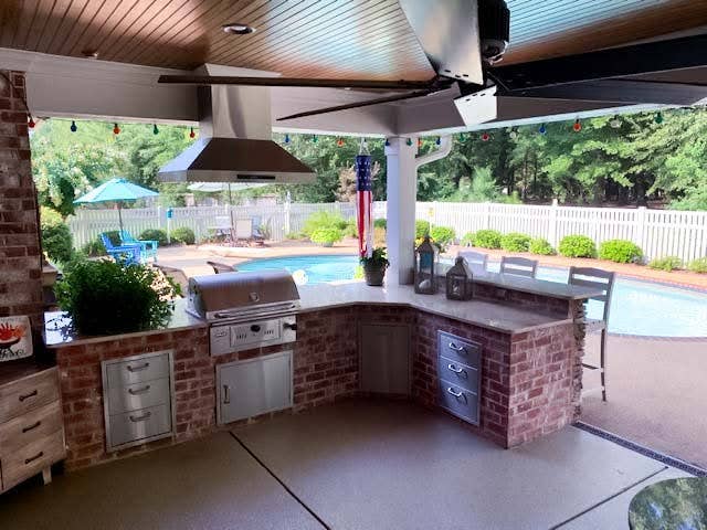 Covered patio with a brick outdoor kitchen featuring a grill and refrigerator, next to a swimming pool with lounge chairs and an American flag. - Proline Range Hoods - prolinerangehoods.com 