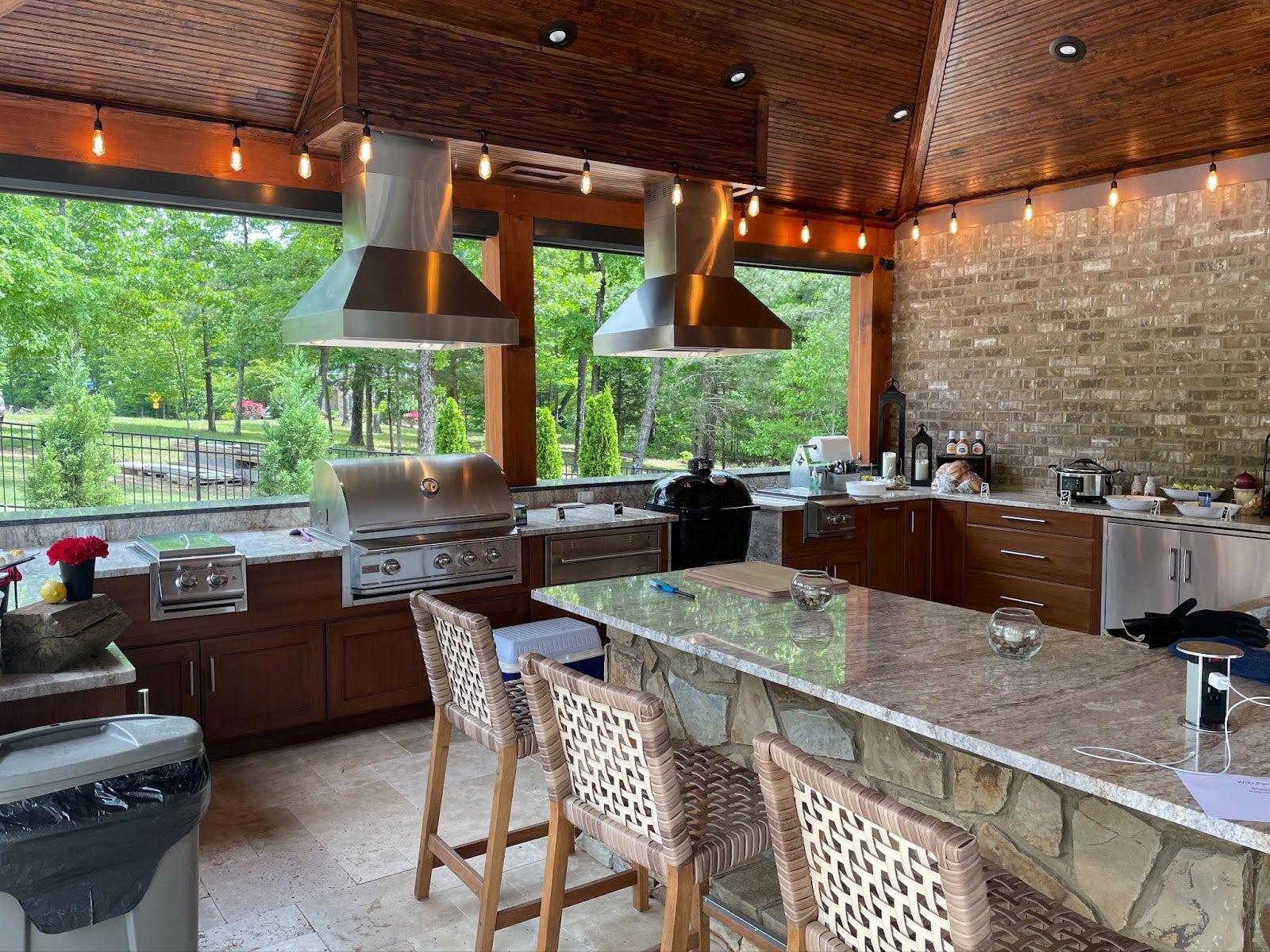 Welcoming open-air cooking space with a natural stone countertop, twin ventilation hoods, and a view of a lush backyard. - Proline Range Hoods - prolinerangehoods.com 