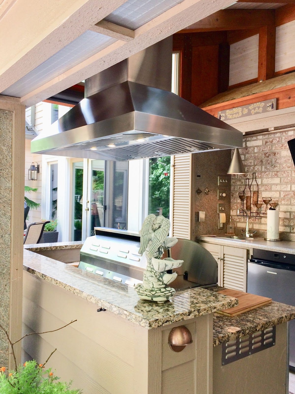 Tranquil backyard cooking nook featuring a modern BBQ grill, vent hood, and a sculpted angel decor on the counter, with a view into the inviting home interior. - Proline Range Hoods - prolinerangehoods.com 
