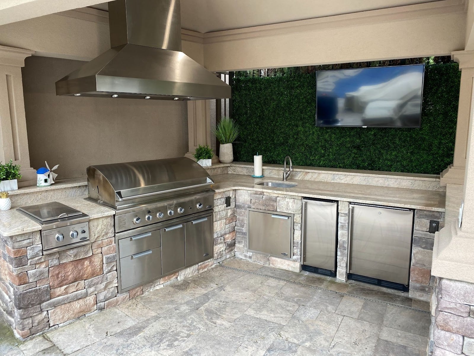 Sleek outdoor cooking area featuring stone worktops, stainless steel appliances, and a mounted television against a lush green backdrop. - Proline Range Hoods - prolinerangehoods.com 