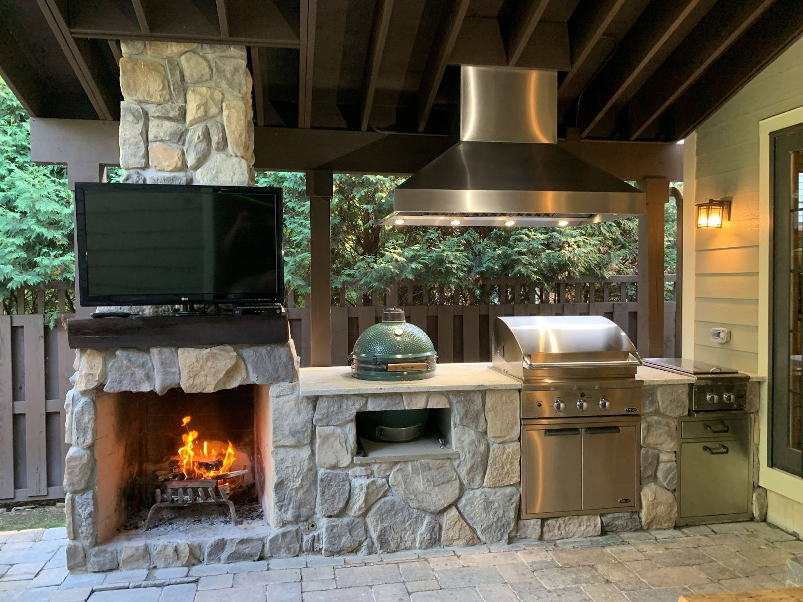 Cozy outdoor patio featuring a stone fireplace with a roaring fire, a modern grill, and a TV for entertainment. - Proline Range Hoods - prolinerangehoods.com 