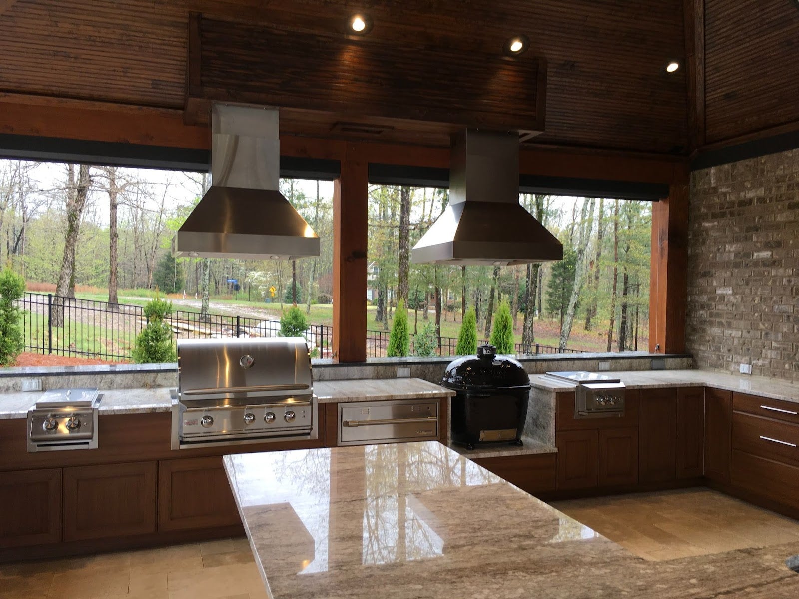 Luxurious and fully-fitted outdoor kitchen with dual extraction units, modern appliances, and wooden storage, surrounded by a tranquil woodland setting. - Proline Range Hoods - prolinerangehoods.com 