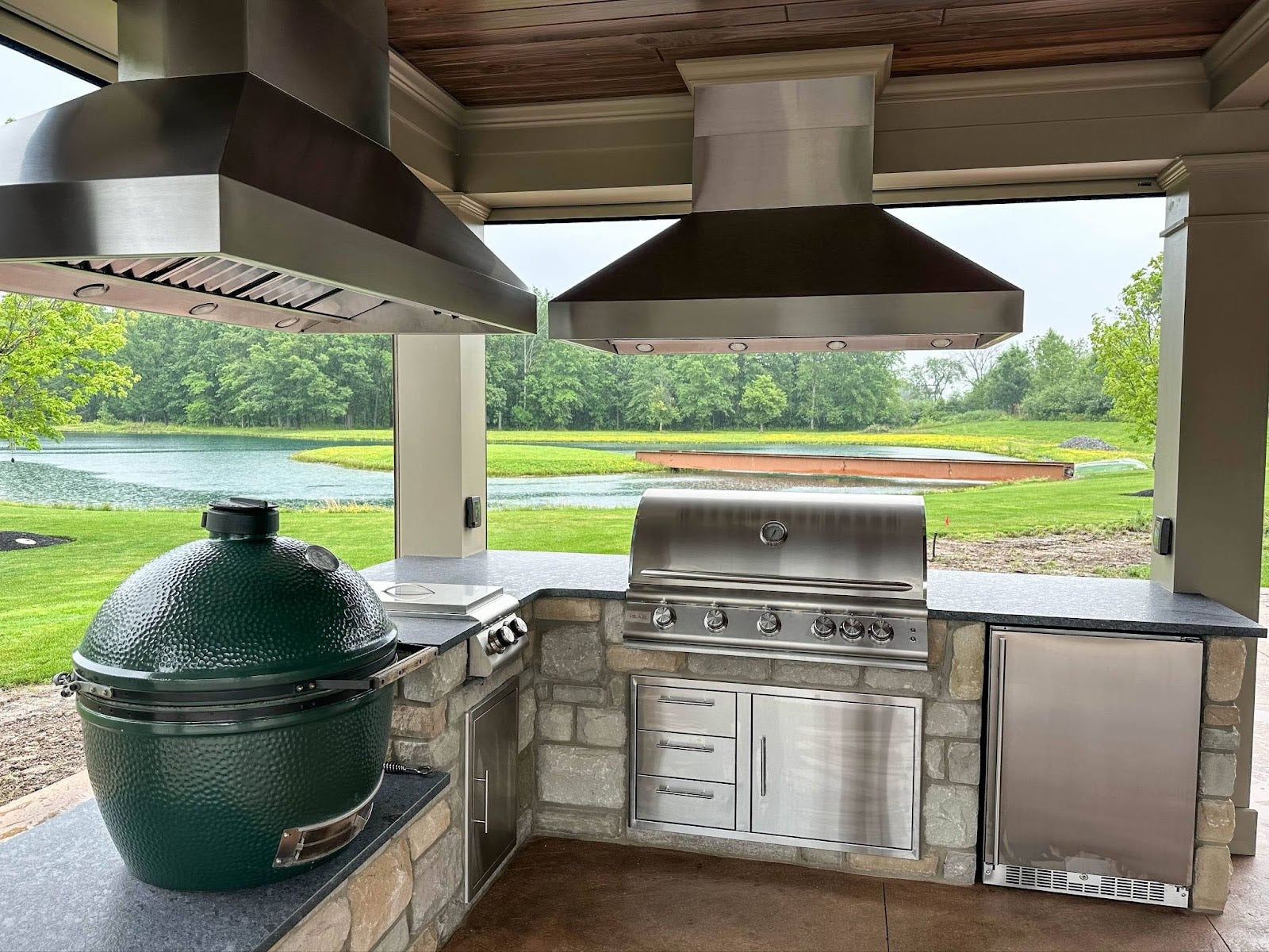 Sophisticated and functional outdoor grilling area equipped with a modern BBQ, green egg smoker, and a refrigeration unit, set against a picturesque pond backdrop. - Proline Range Hoods - prolinerangehoods.com 