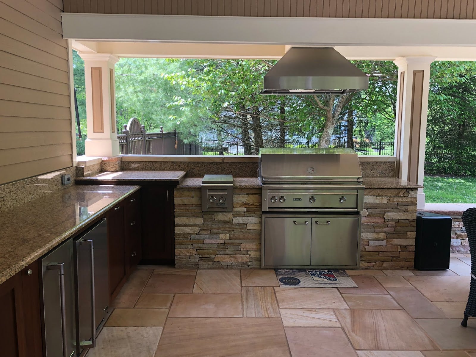 Covered patio kitchen featuring stone accents, modern appliances, and expansive counter space in a serene backyard setting. - Proline Range Hoods - prolinerangehoods.com 