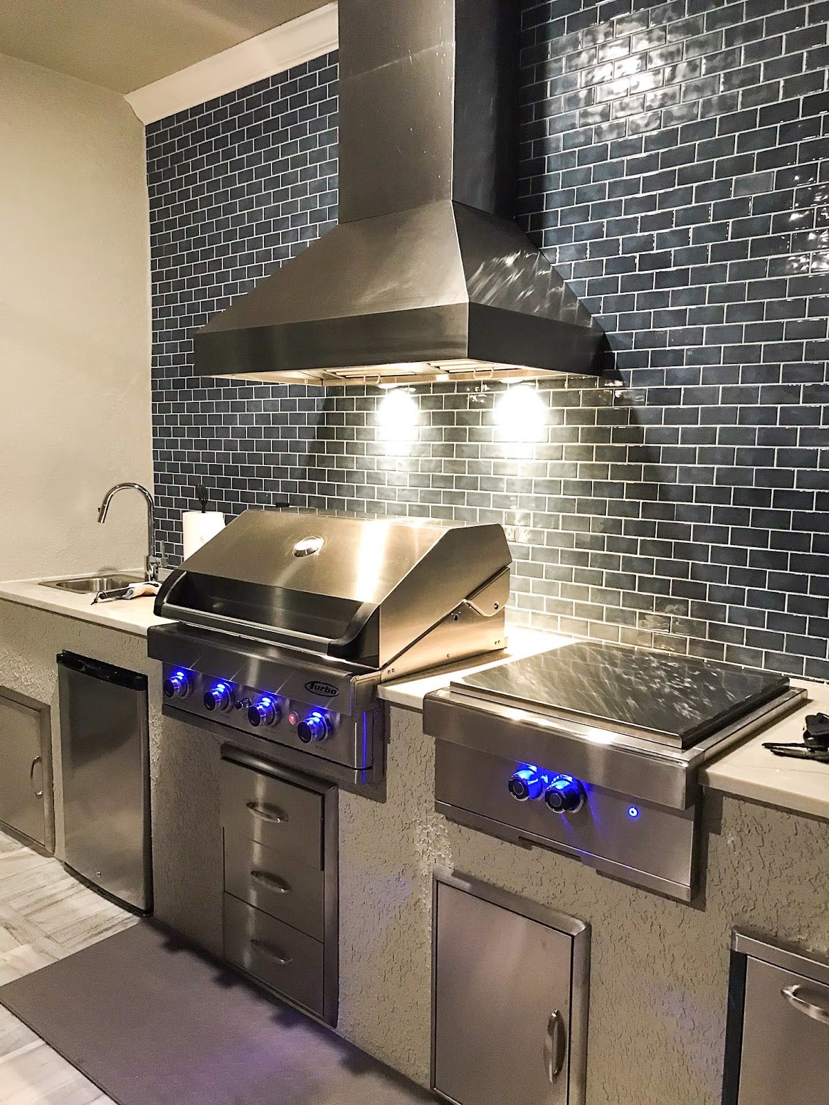Chic outdoor kitchen grill area with a Proline range hood, illuminated by under-hood lighting, against a backdrop of dark subway tiles - prolinerangehoods.com.