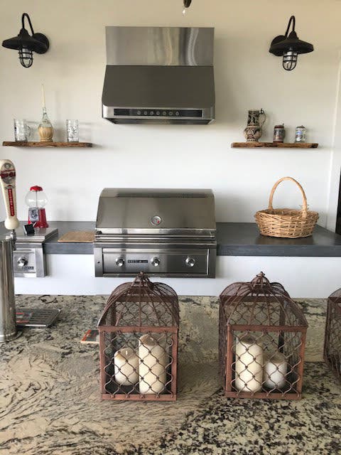 Outdoor kitchen with stainless steel appliances including a grill and range hood, open shelving with decorative jars and baskets, industrial-style sconce lighting, and rustic metal birdcages on the countertop creating a cozy yet modern aesthetic. - prolinerangehoods.com