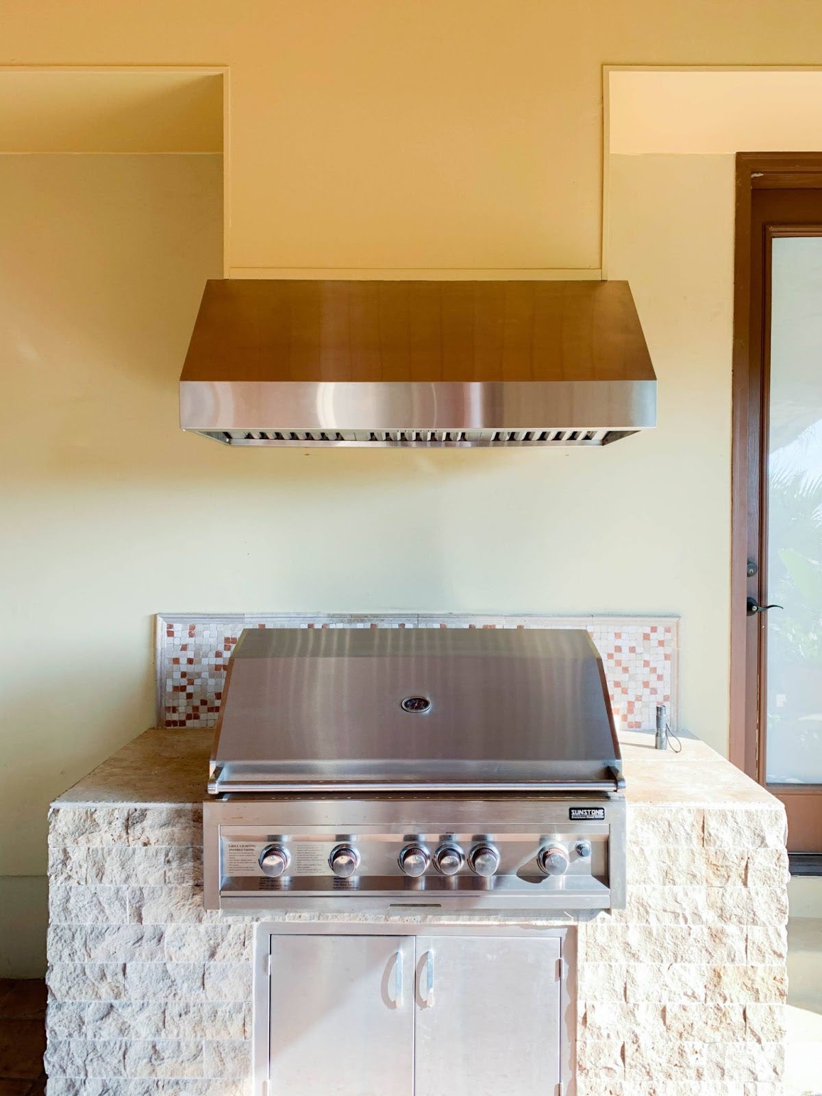 Minimalist outdoor kitchen showcasing a Proline wall mount range hood over a professional grill, complemented by a stone island and mosaic backsplash - prolinerangehoods.com
